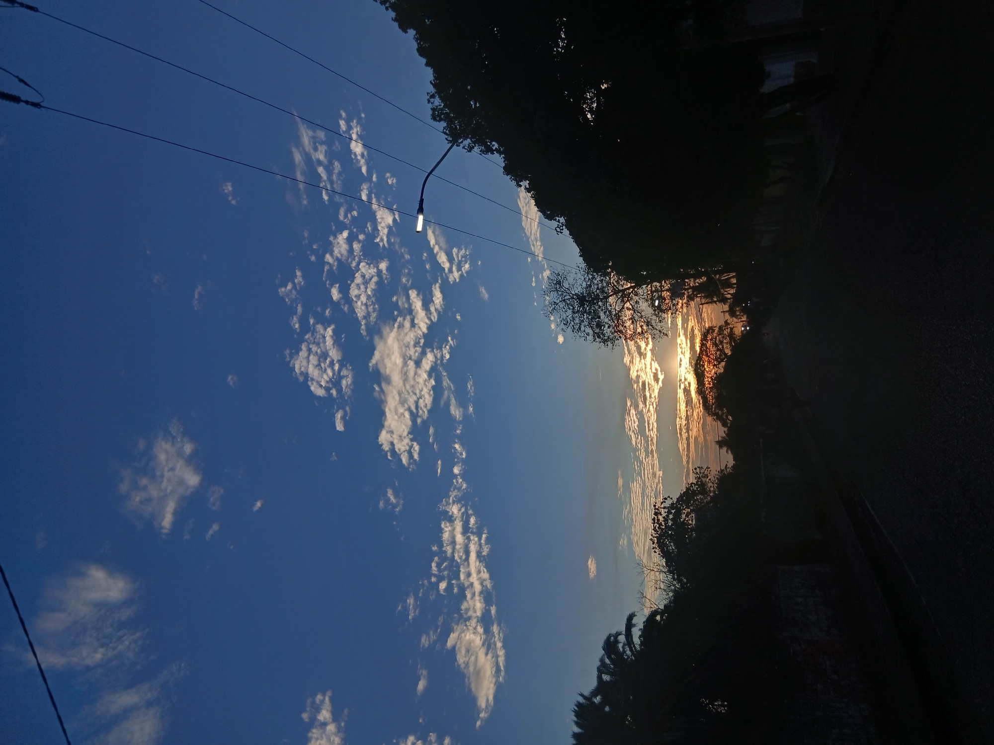 The image shows a view in Juan Griego, Venezuela, at either sunrise or sunset. The sky is blue with scattered white clouds. A streetlamp is visible, hanging from wires extending across the frame. The silhouette of trees and foliage lines the right side of the image, along with a road. The sun is partially obscured by clouds and trees on the horizon, creating a bright, glowing effect. The left side of the image contains clouds and trees.