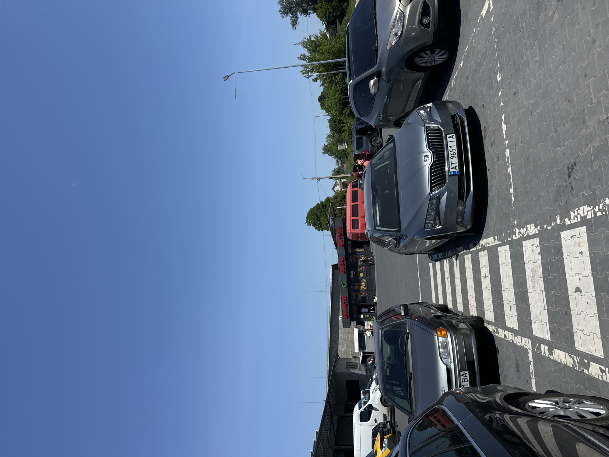 The image captures a bright, sunny day at a parking lot in the city of Sarny, Ukraine. In the foreground, several cars are parked on a gray paved surface marked with white pedestrian crossing lines. A dark gray sedan is visible on the left, partially cropped, with its rear wheel and fender in view. Next to it, a light gray sedan is parked, facing forward, with its license plate readable as "1 BA". Further to the right, a silver Skoda Superb is parked, also facing forward, with its license plate showing "UA AT 9651 IA". Behind it, a silver Hyundai SUV is parked. In the middle ground, a small storefront with a red awning and signage is visible, displaying "ФРУКТИ" (Fruits) and "Овочі" (Vegetables). Several people are seen near the storefront, some appearing to be shoppers. A red mini-bus is parked to the left of the storefront. In the background, industrial-style buildings with gray roofs and concrete block walls are present. Trees and power lines are visible in the distance, under a clear blue sky. The overall scene depicts a commercial area with a parking lot and a small market.