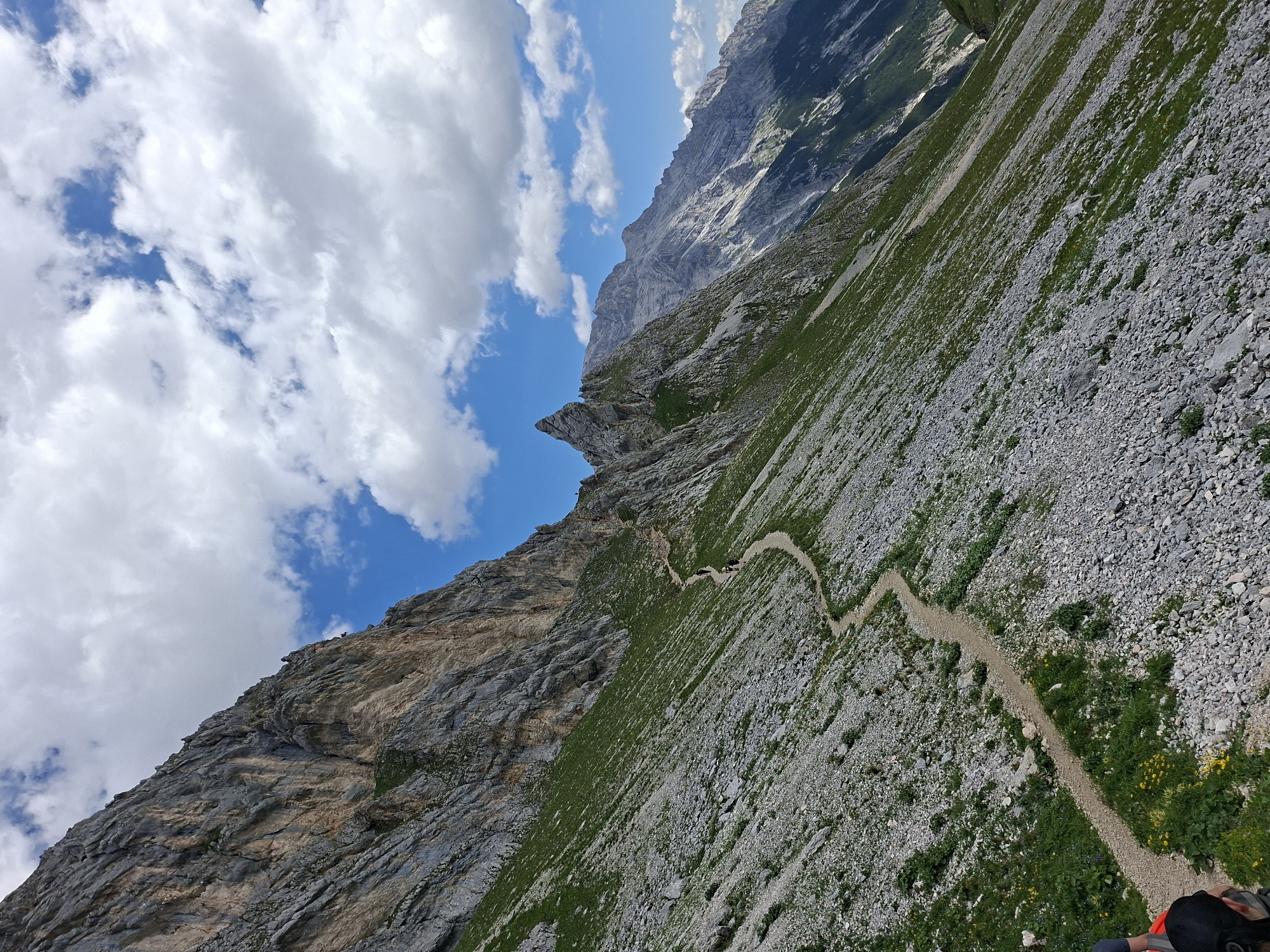 A wide-angle, upward-angled shot captures a rugged mountain trail winding through a steep, rocky slope in the Austrian Alps, near Wildermieming. The path, a light-colored gravel track, snakes its way up the incline, interspersed with patches of green grass and scattered yellow wildflowers. Towering, imposing grey and tan rock faces dominate the left side of the frame, their stratified layers indicative of immense geological age. In the distance, a chain of jagged, grey mountain peaks stretches across the horizon under a brilliant blue sky dotted with fluffy white clouds. Three figures, appearing as small silhouettes, are discernible on the trail in the middle ground, suggesting they are hikers ascending the mountain. The overall impression is one of vast, untamed natural beauty, highlighting the challenging terrain of the Alpine landscape.