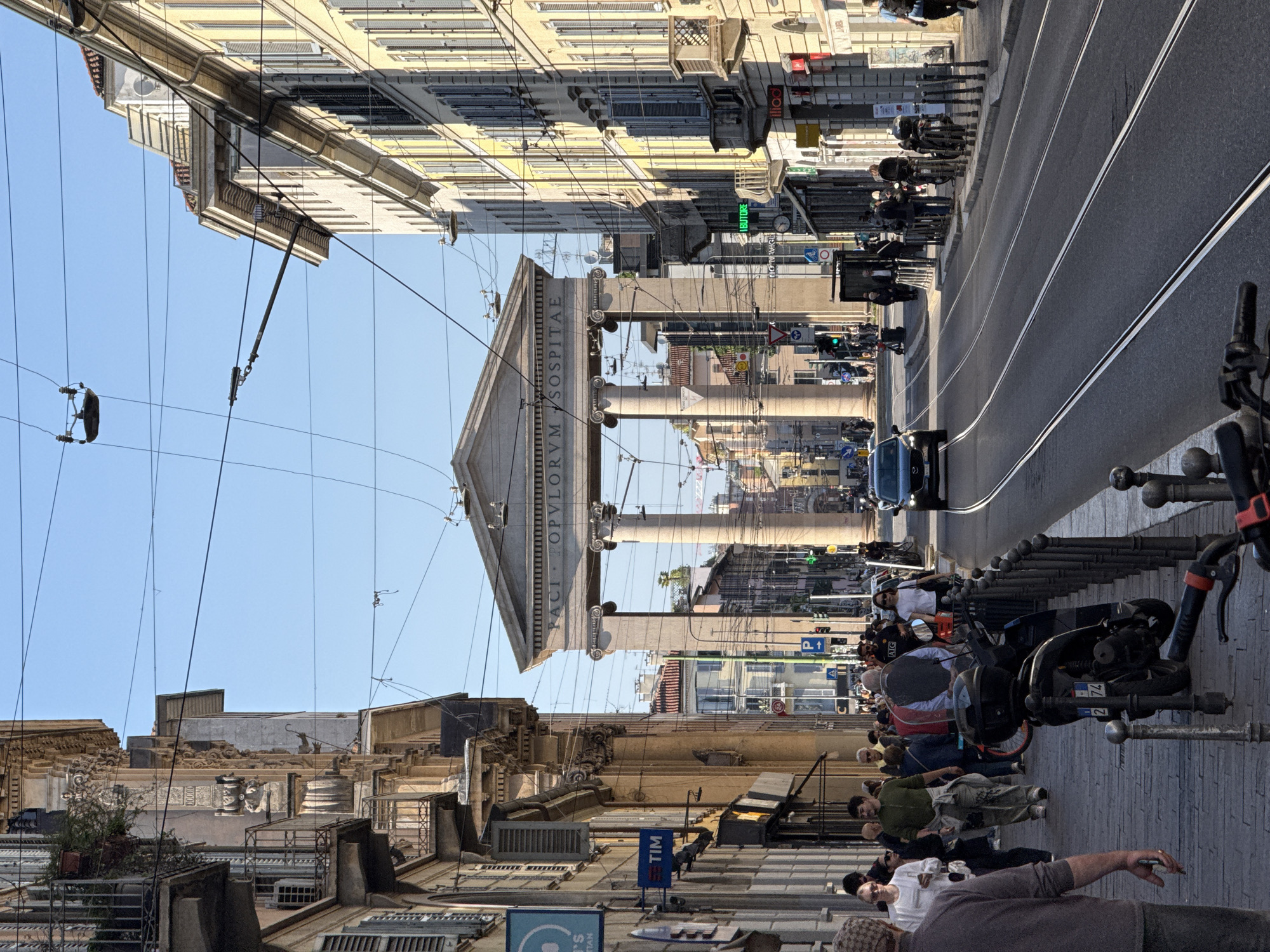 This image captures a bustling street scene in Milan, Italy, under a clear, bright blue sky. The photo appears to have been taken during the day, with bright sunlight casting distinct shadows. The perspective is angled, looking down a street lined with buildings.
In the midground, a prominent neoclassical building dominates the view, featuring large columns and a pediment. Inscribed on the pediment, the text "ORVM HOSPITAE" is clearly visible, part of what is likely "FORVM HOSPITALE" (referring to the historic Ospedale Maggiore, also known as Ca' Granda). Overhead, numerous intricate wires, characteristic of a city with trams or trolleybuses, crisscross the sky.
The street itself, visible in the center and extending towards the foreground, features asphalt and distinct tram tracks running parallel. A dark-colored car is seen driving away from the viewer on the street. Along the right side of the street (foreground), a sidewalk is crowded with parked scooters and motorcycles, secured by a series of bollards. Several pedestrians are scattered across the sidewalk, walking or standing, dressed in casual attire.
Other multi-story buildings, typical of European urban architecture with windows and shutters, line both sides of the street, receding into the background. Visible text on various signs includes the "TIM" logo on a blue sign, a partial sign with "BUTTORE," and another reading "INTERNAZIONALI." A standard "P" parking sign and a green traffic light are also discernible, indicating active urban traffic flow. The overall impression is one of everyday city life in a historic setting.