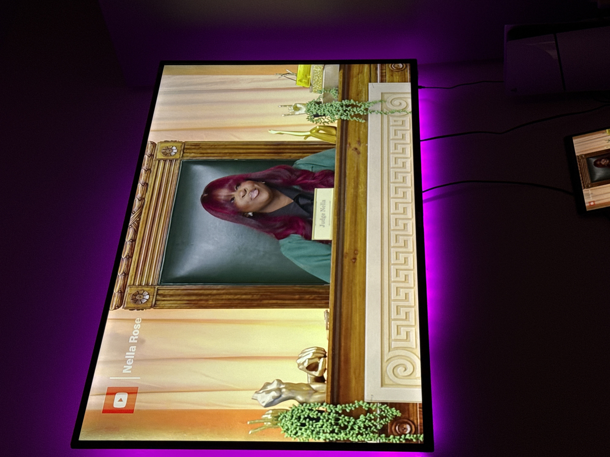 A television screen displays a YouTube video of Judge Nella Rose. The screen is framed with a black border and illuminated with a purple LED strip. The surrounding environment is dark, with a white device visible on the right side of the frame. Additionally, the screen to the right of the monitor is displaying a video call. The location is unknown and is not discernable from the image.