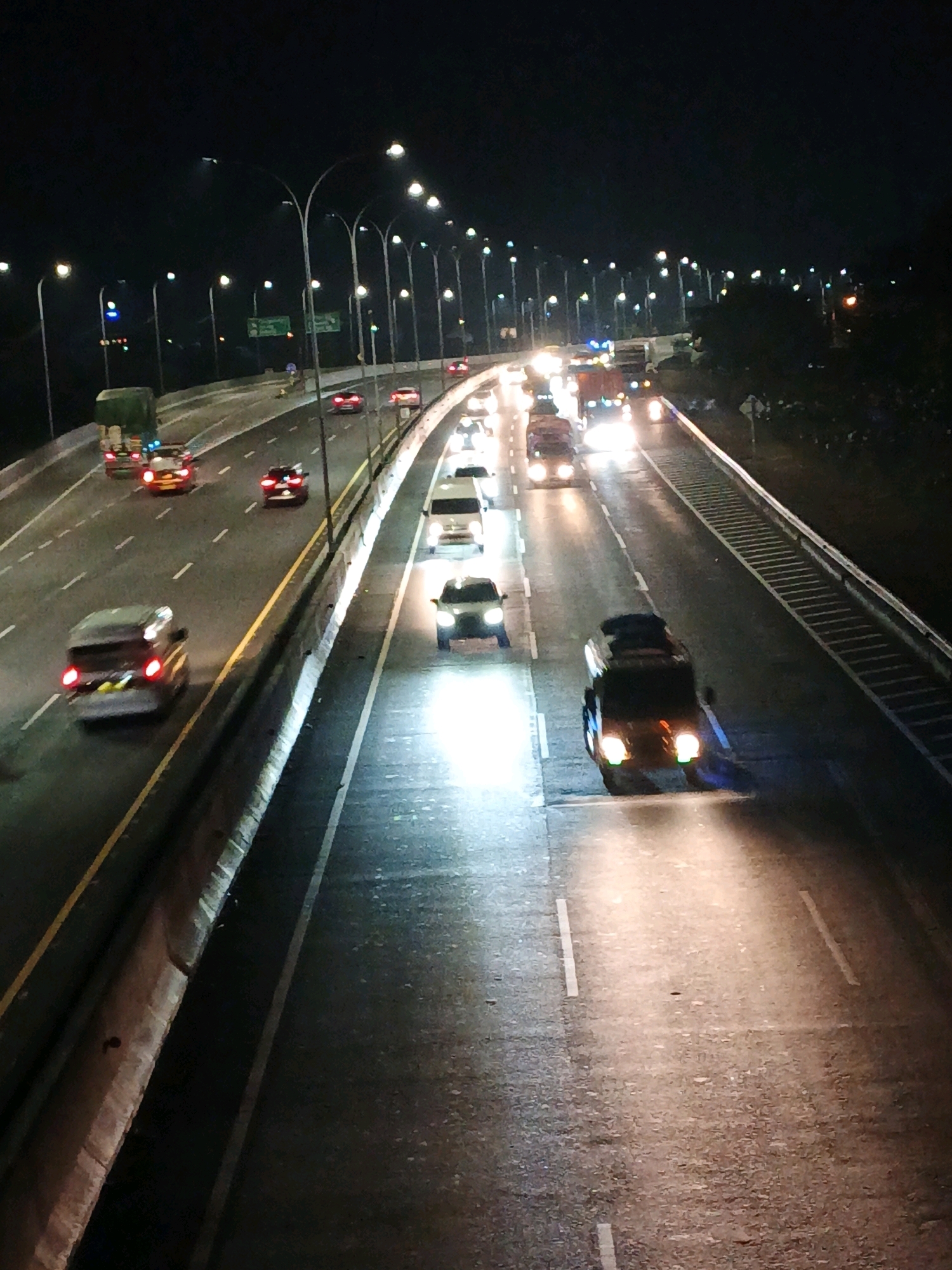 The image depicts a nighttime view of a highway in Kludan, Indonesia, characterized by multiple lanes of traffic illuminated by streetlights and vehicle headlights. Several cars and trucks are visible, with their headlights creating streaks of light on the wet road surface, suggesting recent rain or a damp environment. The overall scene is one of urban transportation at night, with vehicles moving in different directions.