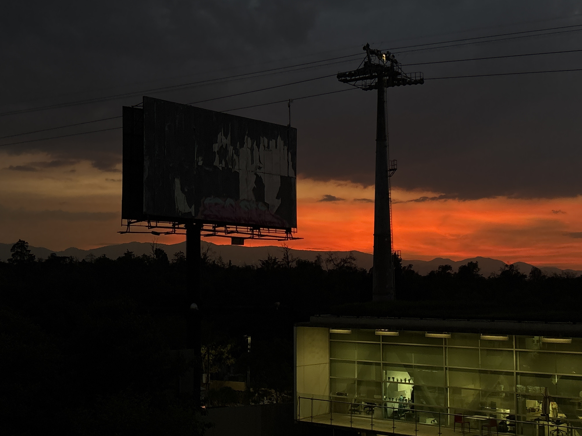 The image depicts a cityscape at dusk in Mexico City, Mexico. The foreground features a modern, low-rise building with large glass windows, illuminated from within. Inside, tables, chairs, and office equipment are visible, suggesting a workspace. A large billboard and a tall metal tower with horizontal supports dominate the midground. Behind these structures, a line of mountains is silhouetted against an orange and red sunset. The sky above the sunset is dark and cloudy. The overall lighting creates a stark contrast between the illuminated building and the silhouettes of the other elements against the colorful sky.