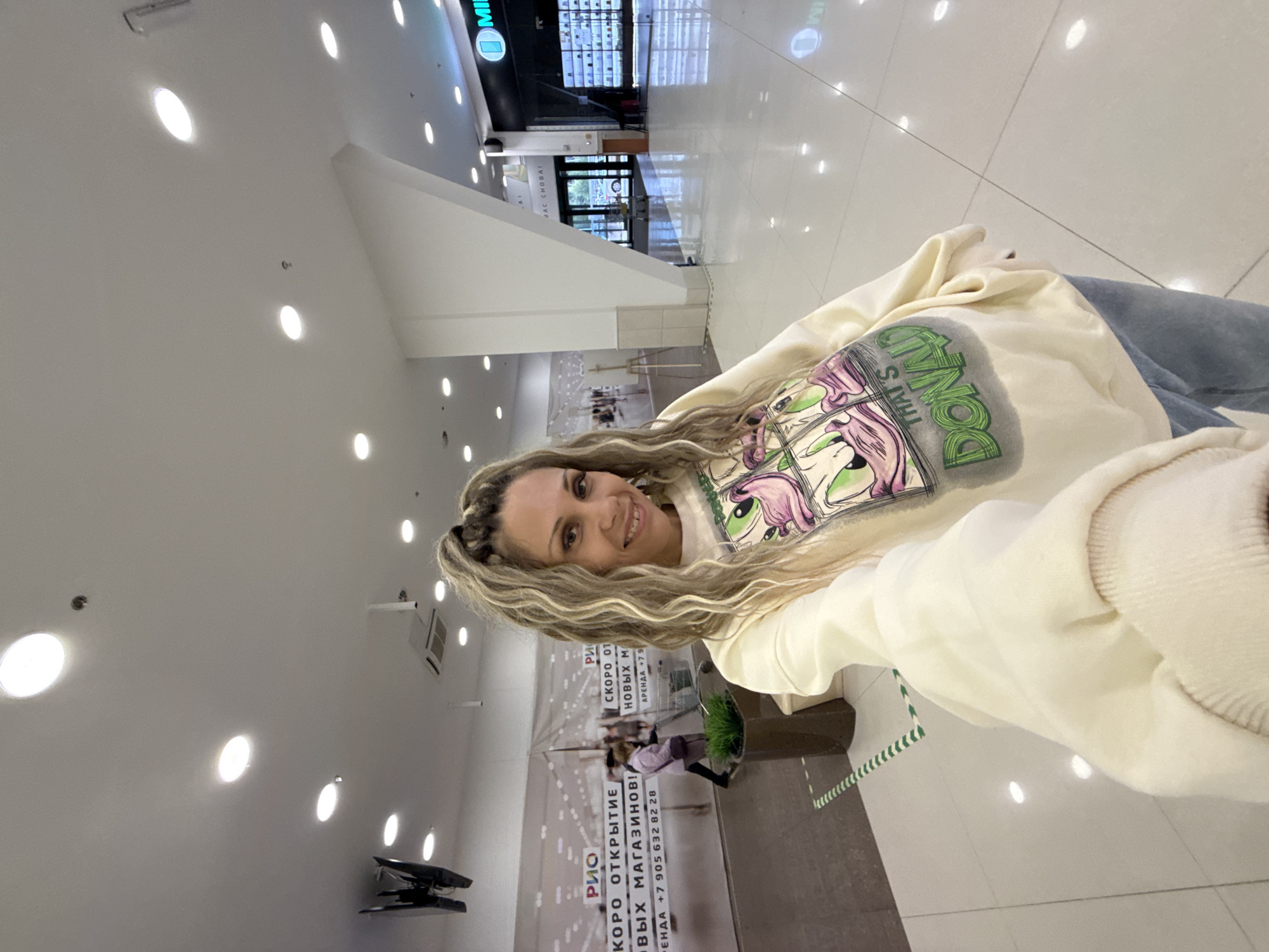 A woman with blonde, braided hair is taking a selfie in what appears to be a shopping mall. She is wearing a light-colored sweatshirt with a graphic of Donald Duck on it and blue jeans. The background shows a bright, well-lit interior with white tiled floors and a white pillar. In the distance, there is a storefront with a sign that says "MI" and displays many shelves of products. To the left of the woman, there is a large banner with Russian text, possibly advertising upcoming openings or store rentals, and a potted plant. Another person is visible in the background, pushing a shopping cart. The overall environment suggests a commercial space.
