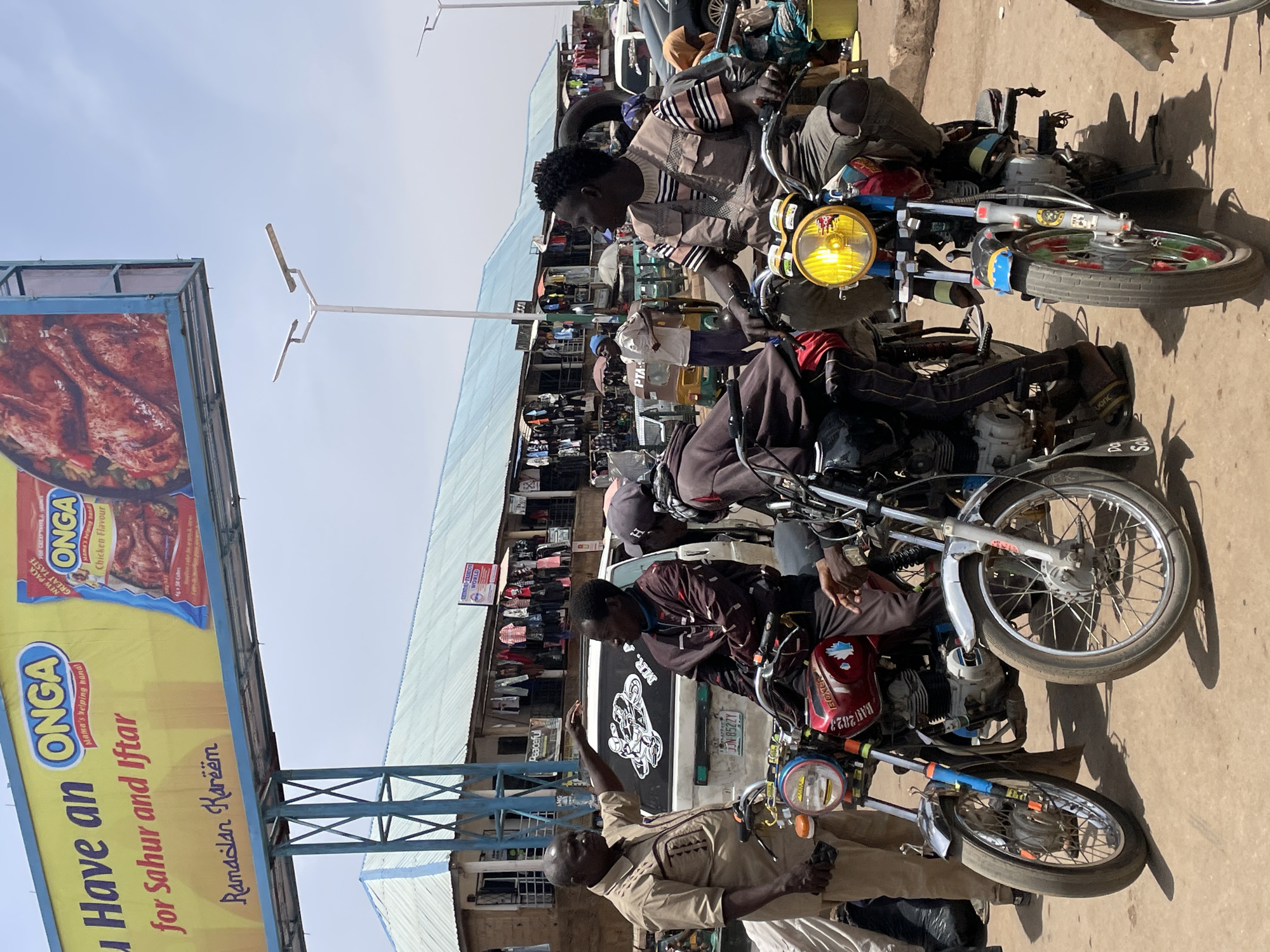 This media file captures a busy daytime street or market scene in Bukuru, Nigeria. The setting features a dirt ground with some paved areas, and various simple buildings with corrugated roofs in the background, suggesting a commercial or residential district. The sky is bright and clear, indicating sunny weather.
The primary subjects are several men and motorcycles. In the foreground, a man in a light brown uniform-like shirt stands facing right, with his left arm raised as if gesturing or directing. Adjacent to him, two men are seated on motorcycles. The man closer to the foreground wears a patterned brown shirt and faces left, while another in a darker shirt is partially obscured. Several other people and motorcycles are visible further back, contributing to the bustling atmosphere. A white van with a black graphic of a figure on a motorcycle is parked behind the men.
The activity appears to be typical market or roadside interaction, with people potentially waiting, conversing, or operating as motorcycle taxi drivers (Okada riders), a common mode of transport in Nigeria. Shadows cast on the ground suggest the sun is high, indicating mid-day.
Notable details include a large yellow and blue billboard on the left, advertising "ONGA" with the text "Have an ONGA Mama's helping hand for Sahur and Iftar Ramadan Kareem." It also mentions "NEW PACK GREAT TASTE" and "ONGA Chicken Flavour." Various other pieces of text are visible on the vehicles and background signs, such as a license plate "JJN 8522Y" on the van, and "BAU2023," "FOXER," "MR. A," "vanc," "Peaceful," "Da," "So" on the motorcycles. A small sign in the background also reads "PTA-". The yellow headlights of the motorcycles stand out against the daytime setting.