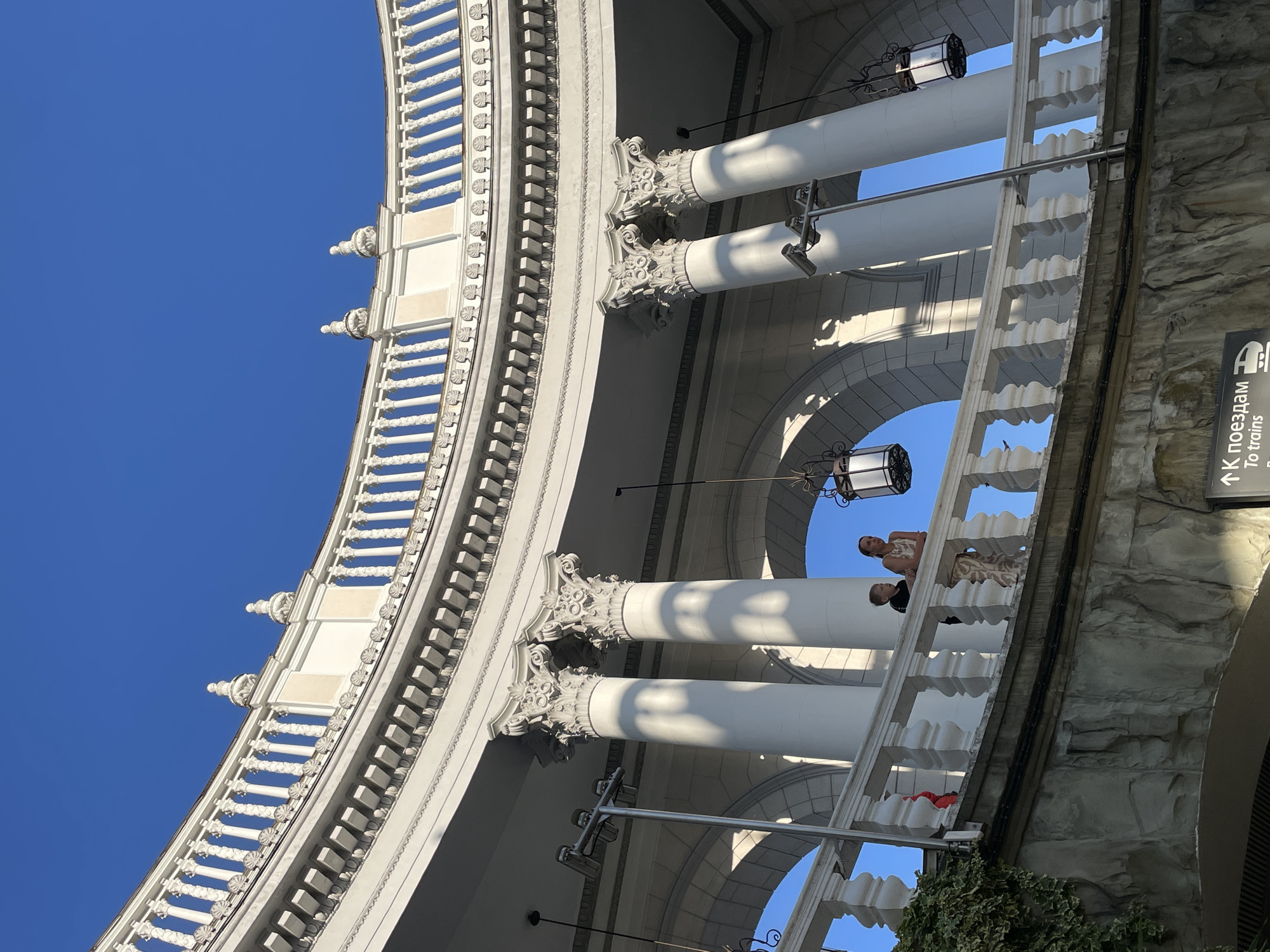 The image shows a curved, white architectural structure with prominent columns and decorative railings against a clear blue sky. The perspective is from below, looking upwards, emphasizing the grandeur of the building. Two people are visible on a balcony or walkway within the structure, appearing to be casually observing the surroundings. A third person, possibly a child, is also present. The lighting suggests it is daytime, likely sunny, with shadows cast by the columns and architectural details.
To the right of the image, a section of the building features a stone-like texture. A sign with text in both Cyrillic and English is partially visible, indicating directions: "К поездам" and "To trains." This, along with the location context provided (Sochi, Russia), suggests the structure might be part of a transportation hub, such as a train station, or a public building in that city. The overall scene conveys a sense of public space and architectural elegance, with the presence of people suggesting a normal day of activity.