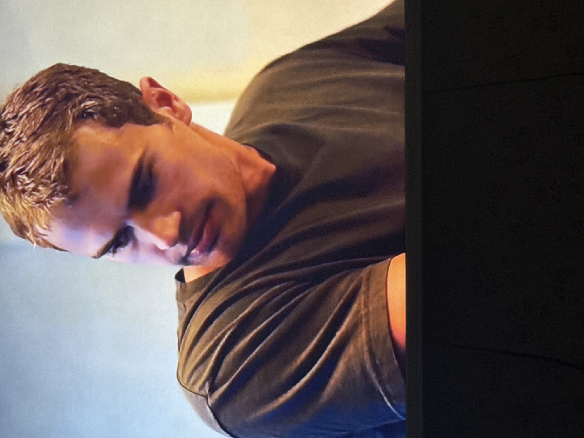 The image shows a man with short, wavy brown hair, wearing a dark-colored t-shirt. His head is turned downward and to the left. The right side of the image is obscured by a dark, undefined object or area. The background appears to be a light-colored or neutral surface. There is no information available from the image about the location in Mytishchi, Russia, or any other information to suggest a specific location or activity.
