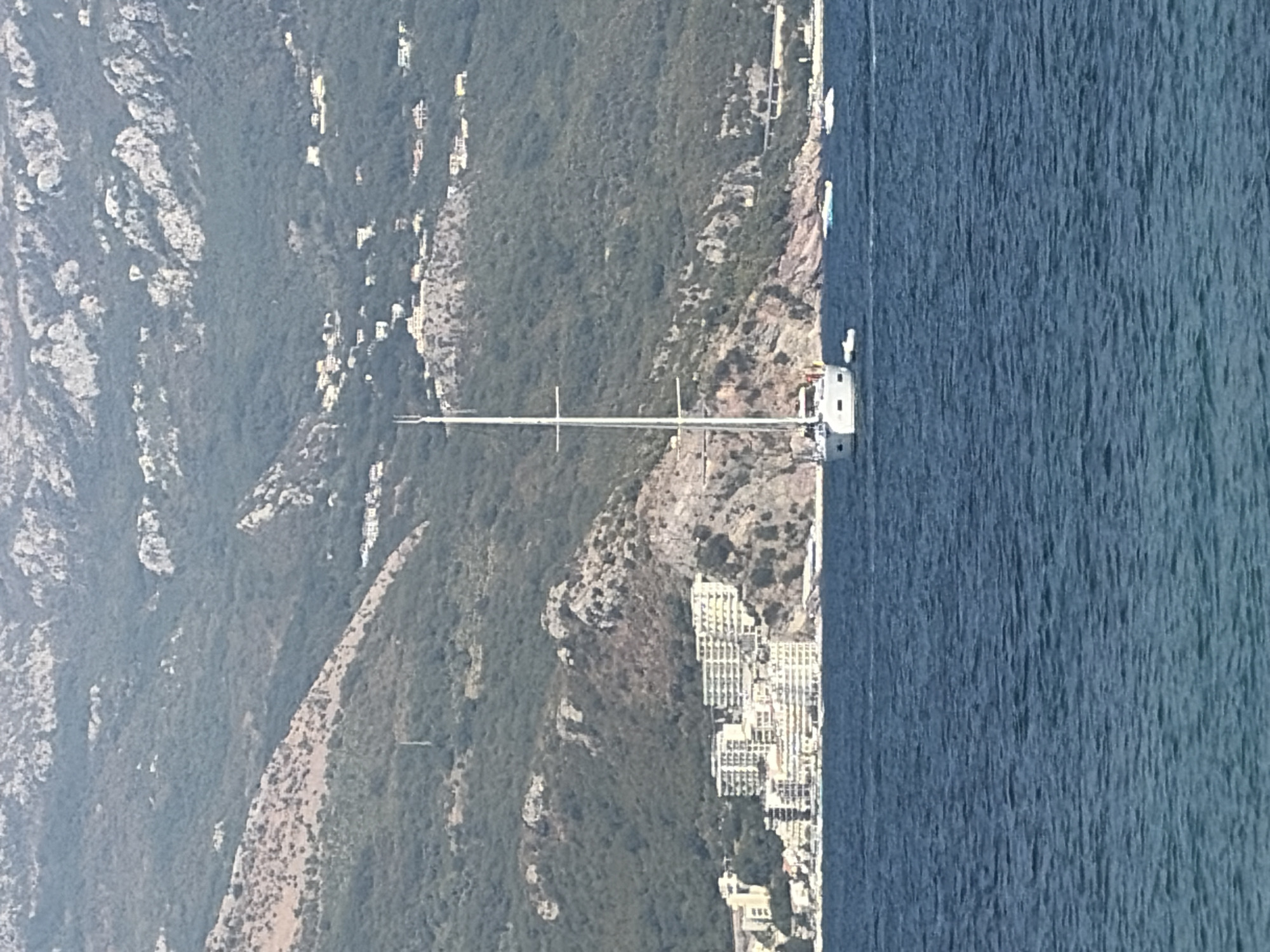 This image captures a scenic view of the Adriatic Sea off the coast of Budva, Montenegro. In the foreground, the deep blue water is calm, with gentle ripples. A white sailboat with a tall mast is the most prominent subject, anchored in the water. Two smaller inflatable watercraft are also visible near the sailboat.
In the mid-ground, a town is nestled at the base of a lush, green, mountainous hillside. The buildings are primarily white, with many balconies, suggesting a residential or hotel area. The architecture appears modern, with several multi-story structures. The hillside is densely covered with trees and vegetation, interspersed with rocky outcrops. Scattered white buildings are visible higher up the slopes, indicating development further into the hills.
The background is dominated by a large, imposing mountain range. The slopes are a mix of dark green vegetation and lighter-colored rocky terrain. The mountains rise sharply, creating a dramatic backdrop to the coastal town. The overall impression is one of a picturesque coastal landscape, with the town seamlessly integrated into the natural environment. The scene appears to be set during daylight hours, with clear skies implied by the brightness of the scene.