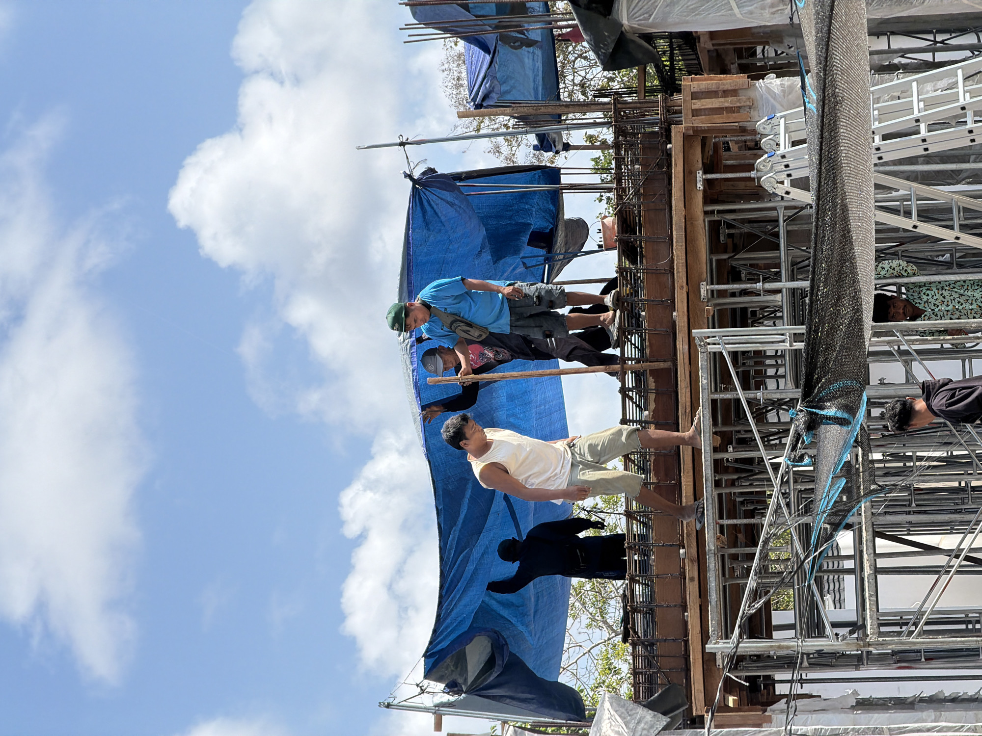 The image depicts a construction site in Kuta Selatan, Indonesia, on a sunny day with scattered clouds. The primary focus is on a group of male construction workers actively engaged in their tasks. In the foreground, a man in a white tank top and khaki shorts walks to the left, appearing to be moving around the elevated platform. To his right, another worker wearing a blue t-shirt and shorts stands, holding a pole, possibly assisting with the blue tarp overhead. Behind them, a third worker in a black shirt and cap is partially visible, also interacting with the tarp.
The construction area features scaffolding, wooden beams, and rebar, indicating ongoing structural work. A large blue tarp is suspended above the workers, providing shade. The environment suggests an outdoor construction project, with trees visible in the background. The overall scene captures the labor and activity characteristic of a building site in progress.