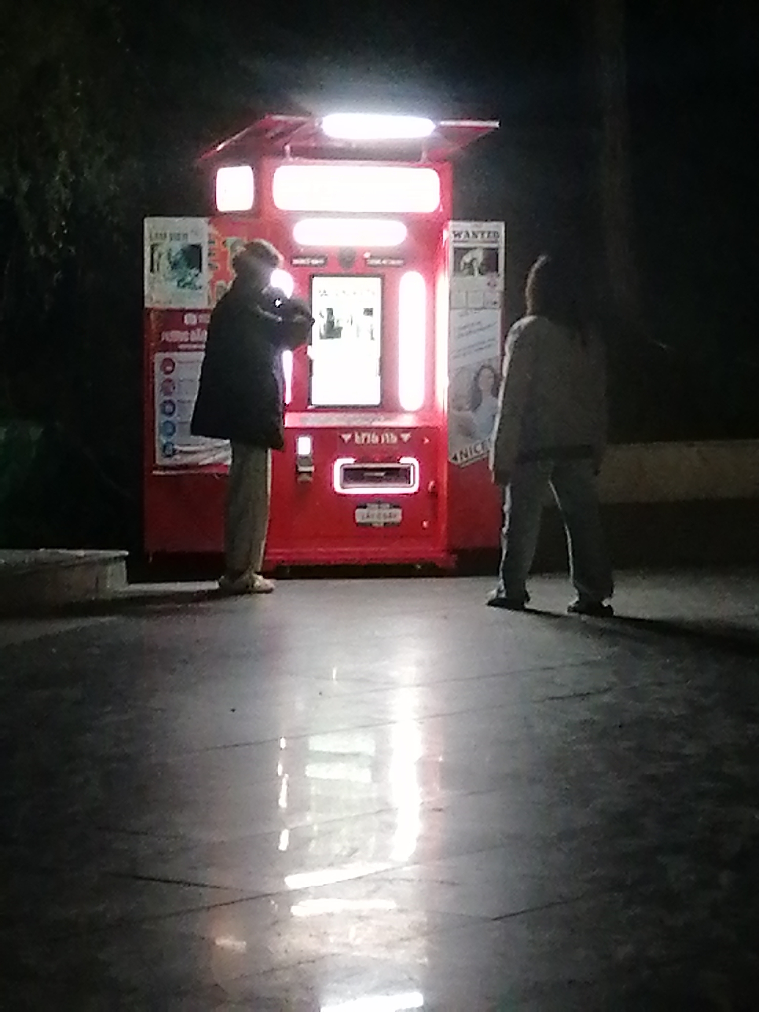 A medium-angle shot captures two people interacting with a red vending machine. The scene is dimly lit, suggesting nighttime. On the left, an individual in a dark jacket and light-colored pants appears to be using the vending machine. To the right, another person in a light-colored jacket and pants stands facing the machine. The machine itself is brightly illuminated and adorned with posters and vending options. The smooth surface in the foreground reflects the light, adding to the overall visibility in the low-light setting. The location is identified as Da Lat, Vietnam.