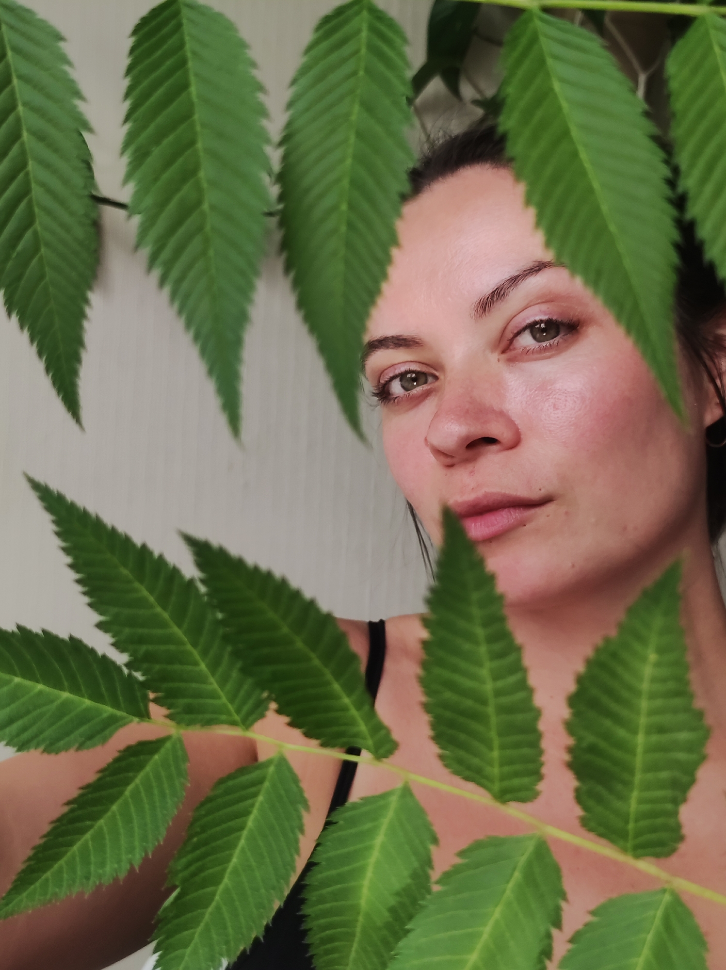 A woman poses close to the camera, her face partially obscured by a leafy green plant. The plant's leaves are pinnately compound, and several stems are visible, creating a frame around her face. Her eyes are green, and her hair is pulled back. She wears a black top, and a white wall is in the background. The image was taken in the city of Chaikovsky, Russia.