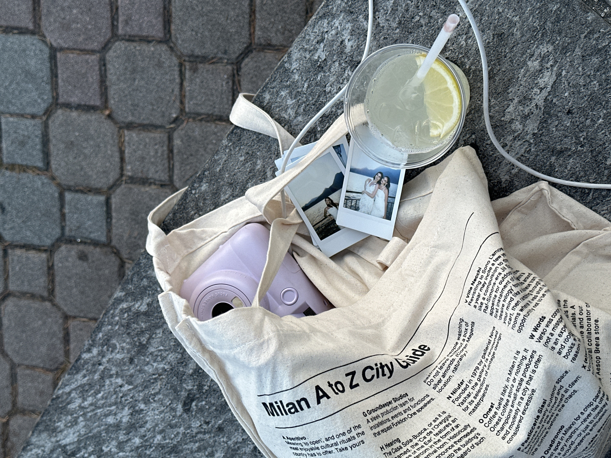 An overhead shot shows a beige tote bag resting on a weathered gray stone surface, possibly a ledge. Inside the bag, among the folds of fabric, is a lavender instant camera, two small instant photos, and the corner of what appears to be a travel guide ("Milan A to Z City Guide"). A clear plastic cup filled with a clear liquid, possibly lemonade, with a straw and a lemon slice sits on top of the bag, along with a white charger cable. The setting appears to be outdoors, with cobblestones visible to the side of the ledge. Gittana, Italy is not mentioned in the text of the bag.