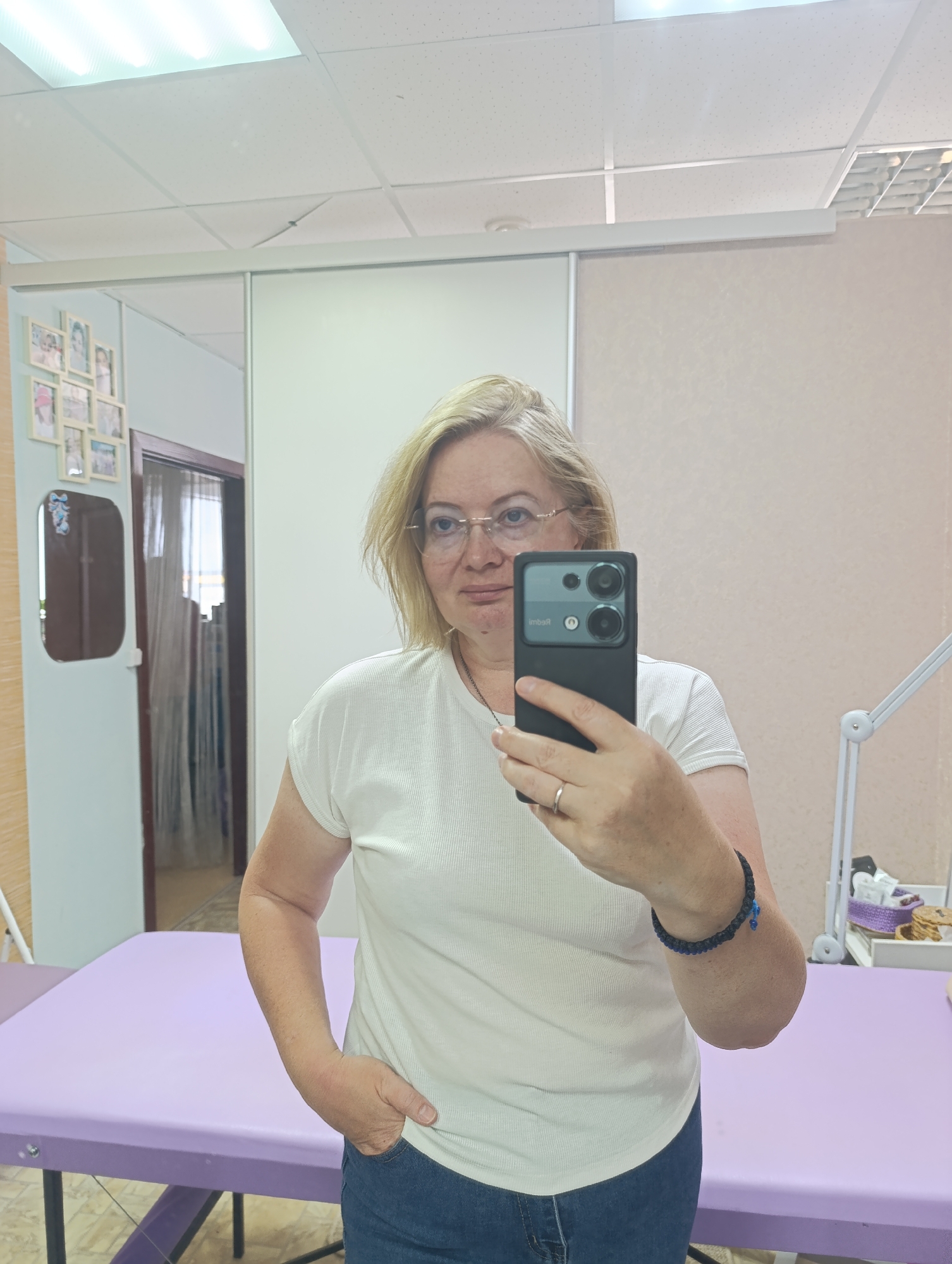 A blonde woman, appearing to be in her 40s or 50s and wearing glasses, a white ribbed t-shirt, and blue jeans, is depicted taking a mirror selfie indoors in Chaikovsky, Russia. She holds a black smartphone with multiple camera lenses in her right hand, while her left hand rests in her pocket. A silver wedding ring is visible on her left ring finger, and a dark blue beaded bracelet with a small blue charm is on her right wrist. The brand name "Redmi" is faintly visible (reversed) on the back of the phone.
The setting is a professional room, likely a beauty salon, clinic, or massage parlor. A prominent feature is a light purple examination or massage table in the foreground. In the background, to the right, a white medical or esthetic lamp with articulated arms is visible, along with a white plastic basket containing various small containers. The room has light-colored walls, with white sliding doors or partitions in the center-right. To the left, a collection of small picture frames and a small, irregular-shaped mirror adorn the wall, next to a door frame with dark brown trim. The ceiling features tiled drop panels with bright fluorescent lighting, suggesting daytime. The woman has a neutral to slightly pleasant expression.