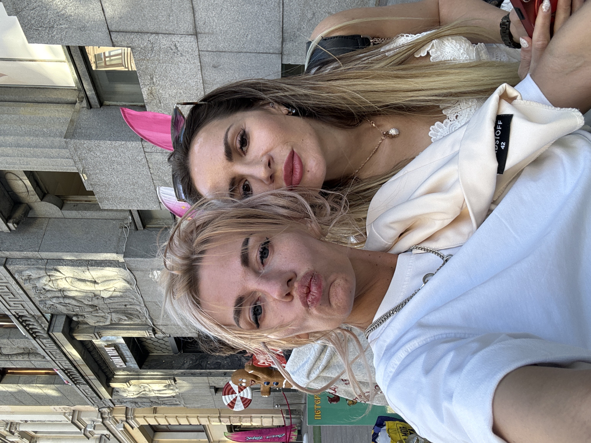 Two women are taking a selfie in front of a building in Saint Petersburg, Russia. The woman on the left has blonde hair and is wearing a white t-shirt and a silver necklace. She is puckering her lips. The woman on the right has long brown hair and is wearing a white lace top and pink bunny ears. She is wearing sunglasses on her head and a necklace with a pendant. The building behind them is made of stone and has ornate carvings. A sign on the building reads "СМЕНА ПАРК". In the background, there are some decorations, including a gingerbread man and a large candy cane. There are also some shopping bags visible.