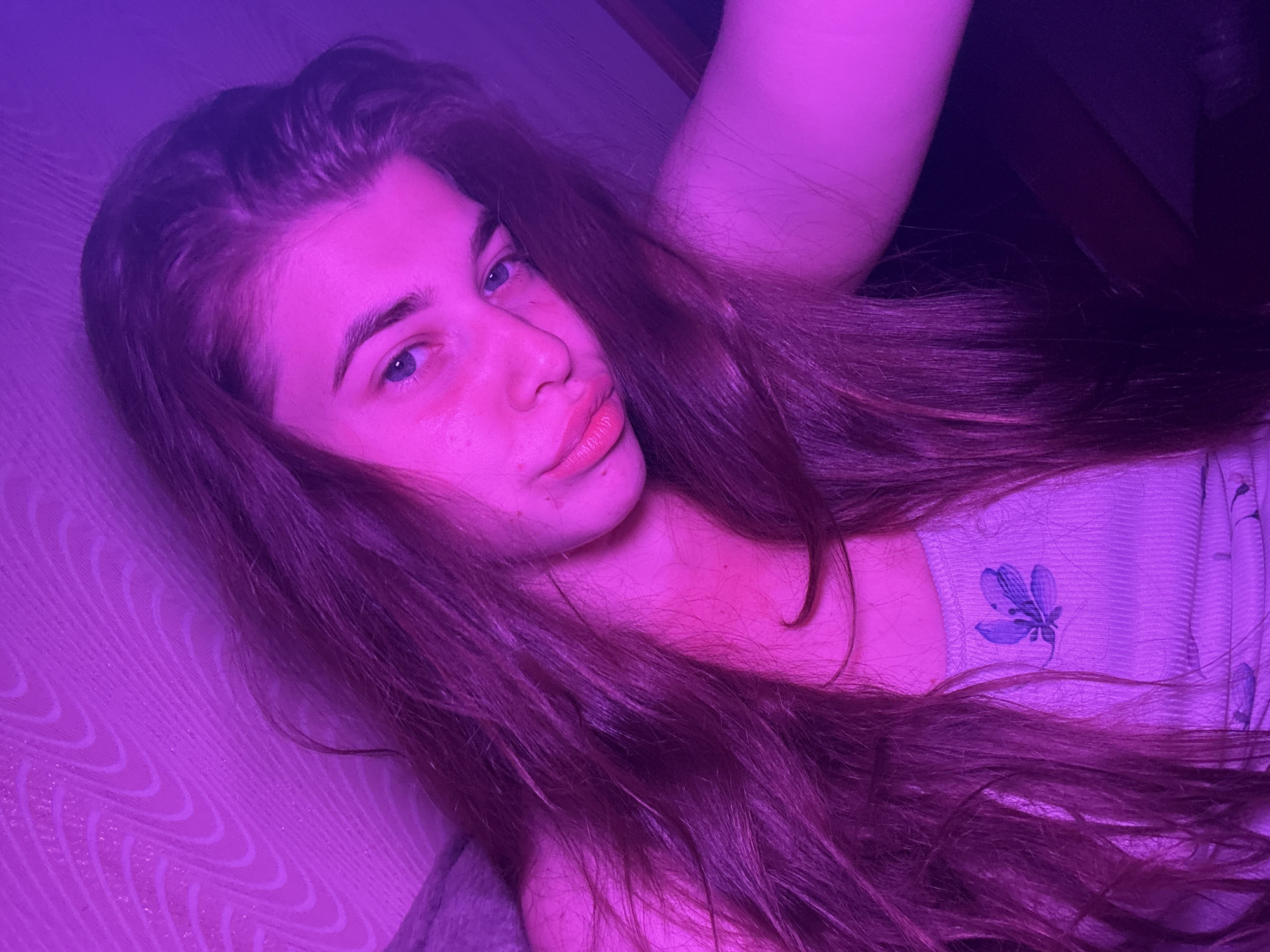A young woman with long, dark hair is pictured in a dimly lit room with purple lighting. She is looking towards the camera with a neutral expression. Her hair is falling around her face and shoulders, partially obscuring her body. She is wearing a light-colored tank top with a floral design. The background appears to be a wall with a textured pattern. There is no information available about the location in the city of Мещёры, Russia.
