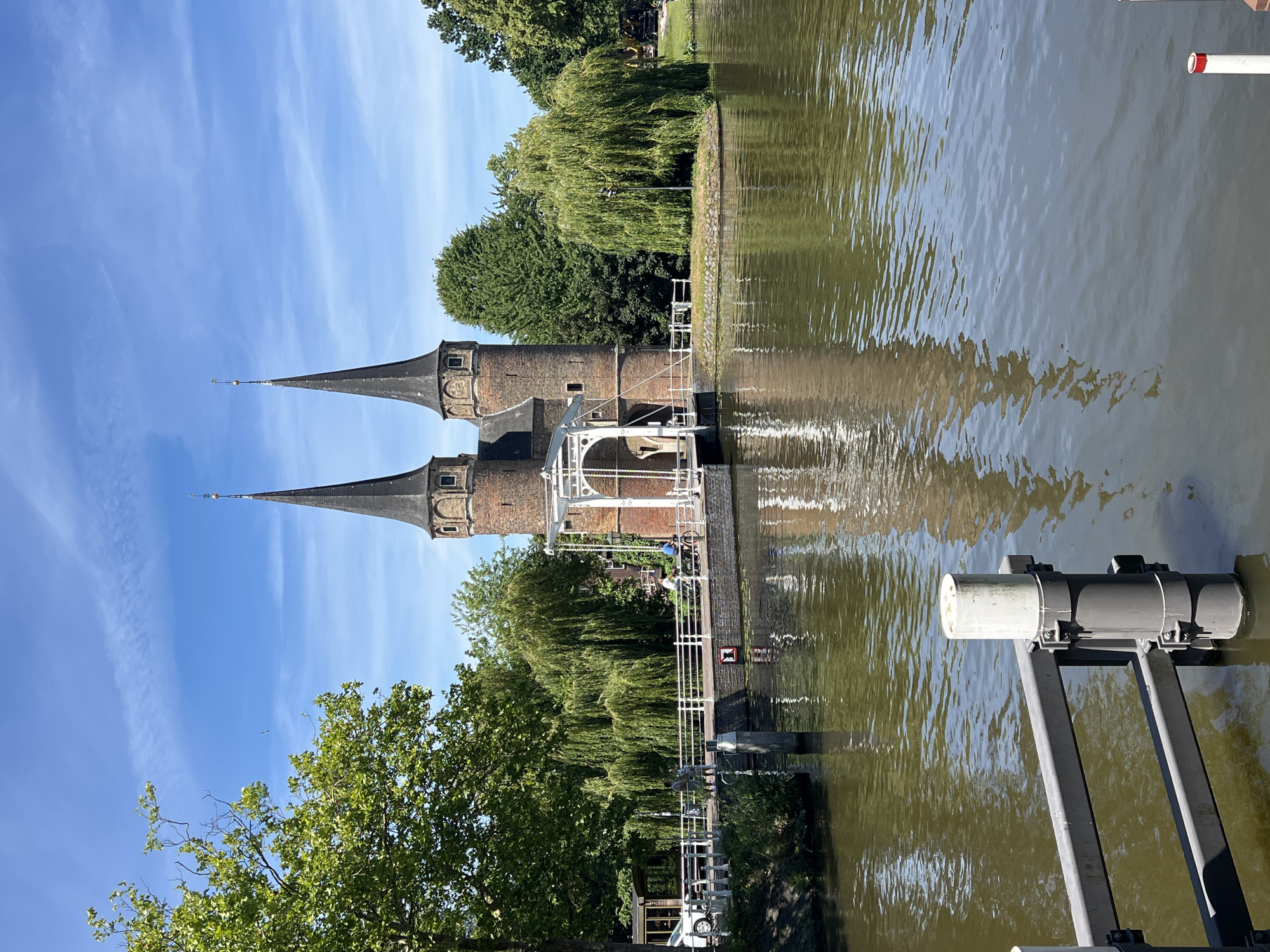 The image shows the Oostpoort, a medieval city gate in Delft, Netherlands. The gate features two conical towers with dark roofs and a white drawbridge in the center. The gate is made of red brick and has several windows. The Oostpoort is situated on a canal, with its reflection visible in the water. Surrounding the gate are lush green trees, including several weeping willows. On the bridge, there are a few people, some walking and others cycling. To the left of the bridge, a white car is parked. The sky is a clear blue with some wispy clouds. The water in the canal is calm, with ripples on the surface. In the foreground, a section of a metal railing and a white post are visible, suggesting a dock or a pier. The overall scene depicts a sunny day in a picturesque historical setting.