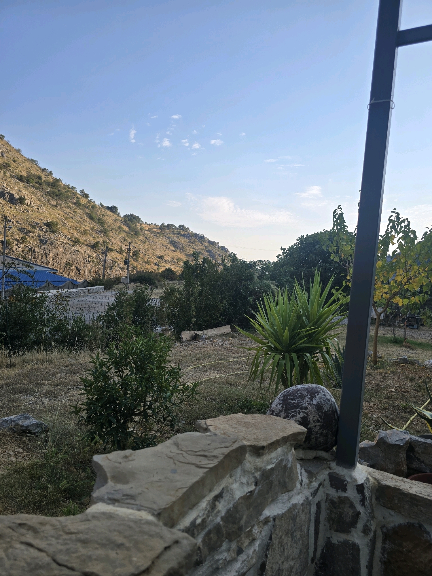 The image depicts an outdoor scene with a dry, rocky hillside in the background under a clear blue sky with sparse white clouds. In the foreground, a low stone wall is visible, constructed from irregularly shaped rocks and mortar. To the right of the wall, a dark vertical metal pole rises upwards. Beyond the wall, the landscape features dry grass, a spiky green plant resembling a yucca, and a small tree with yellowing leaves. A larger, rough-textured grey spherical object rests on the stone wall behind the spiky plant. In the mid-ground, there is a chain-link fence and a blue tarpaulin covering a structure. Power lines and poles are also visible on the hillside. The overall impression is of a rural or semi-rural setting, possibly in a Mediterranean climate, given the vegetation and dry landscape. The location is identified as Podgorica, Montenegro. There are no people visible in the image.