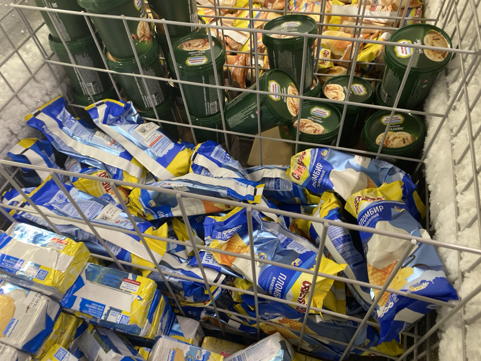 The image shows a close-up, eye-level view of a wire freezer bin filled with frozen food items. The bin is packed with a variety of individually wrapped ice cream products, many of which have blue and yellow packaging. There are also several green cylindrical containers stacked on shelves within the bin. The lighting is dim and appears to be artificial, likely from within the freezer. The overall scene suggests a commercial setting such as a grocery store or supermarket freezer aisle. The location is unknown.