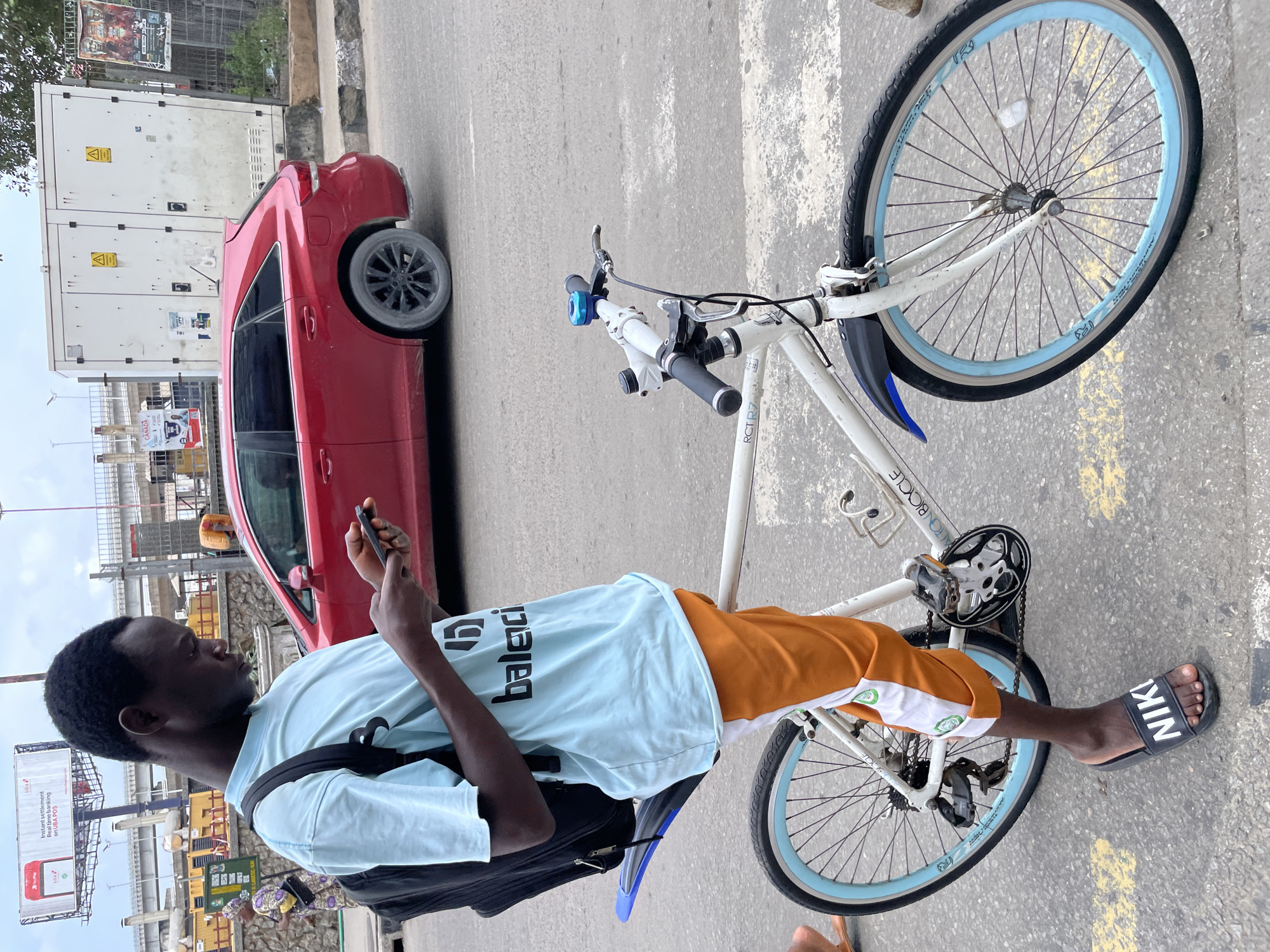 A young Black male wearing a light blue t-shirt with black lettering and orange shorts is standing next to a white bicycle. He is looking down at his phone, which he holds in his right hand. The bicycle has a black backpack strapped to the handlebars and a blue fender over the front wheel. The bicycle also has light blue rims on both wheels. The scene is outdoors in what appears to be an urban environment. In the background, a red car is parked on the street, and a large white electrical transformer box is visible. The ground is paved with asphalt and marked with yellow lines, suggesting a street or sidewalk. There is a billboard in the distance that appears to be an advertisement. The overall lighting suggests an overcast day. The man is wearing black Nike slides on his feet.