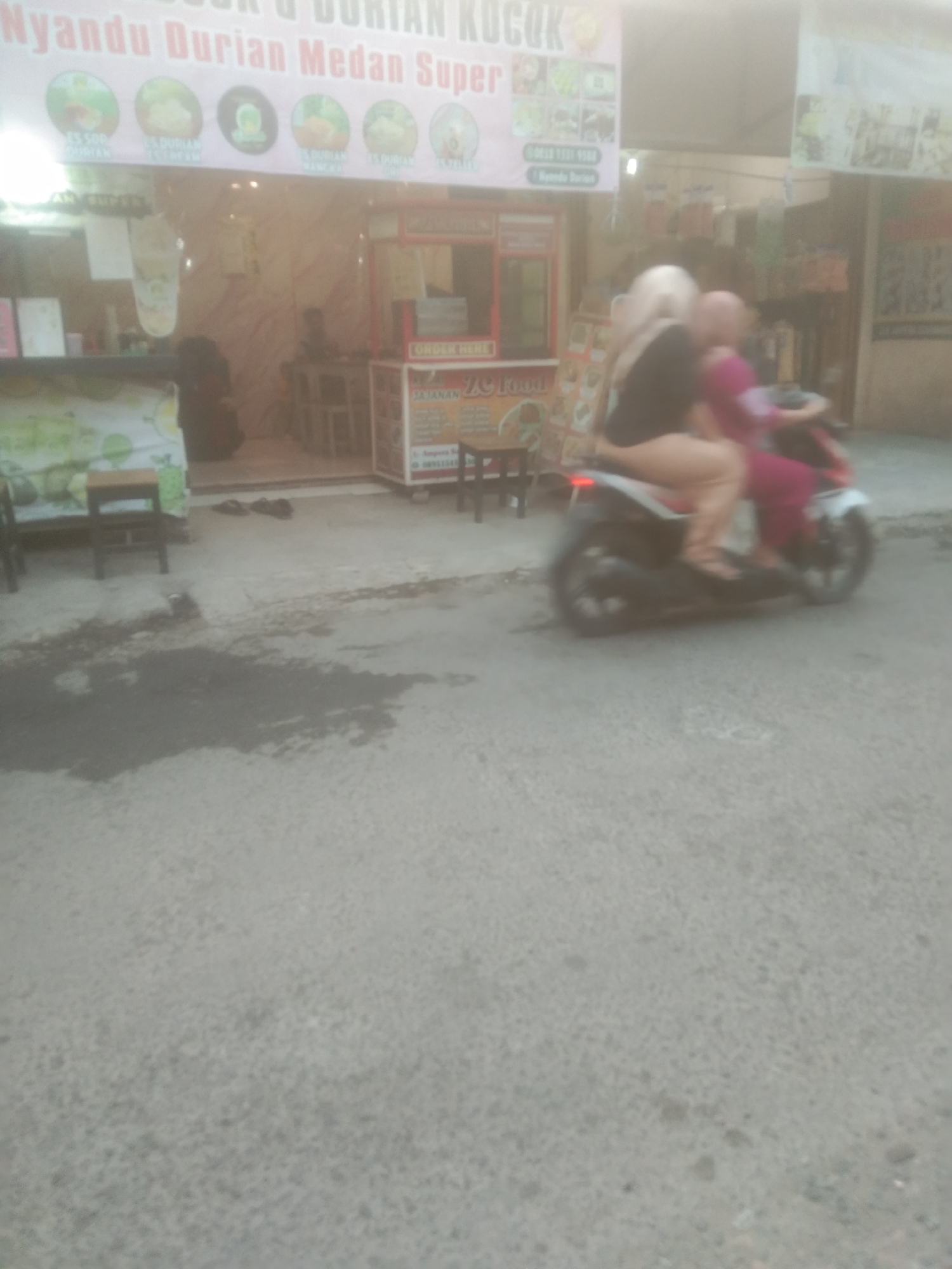 The image depicts a street scene in Subang, Indonesia, with a focus on a small shop or food stall. A sign with the text "Nyandu Durian Medan Super" is visible above the shop. The shop appears to be selling durian products. In the foreground, a motorcycle is seen moving, carrying two passengers. The woman in the front is wearing a black top and beige pants, while the woman behind her is wearing a pink outfit. The street is paved and appears to be wet in some areas, possibly from rain. Several chairs are visible outside the shop, suggesting a seating area for customers. The overall atmosphere is that of a typical urban street in Indonesia, with bustling activity.