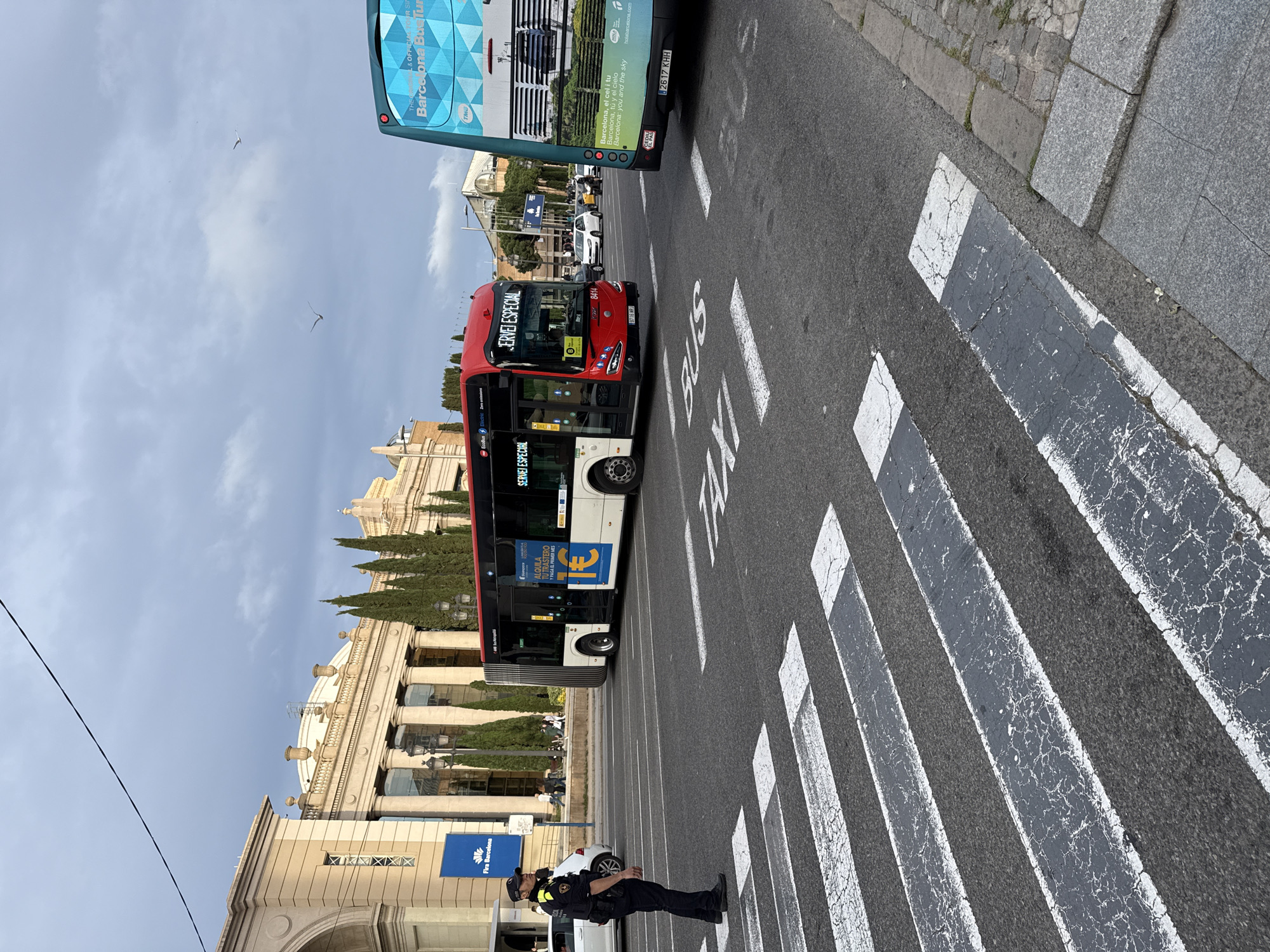 The image shows a street scene in Barcelona, Spain. A red and white "Servei Especial" bus is crossing a pedestrian crosswalk with "BUS" and "TAXI" painted on the road. A police officer in a dark uniform stands near the crosswalk, presumably directing traffic. In the background, a large, ornate building is visible, which appears to be the Fira Barcelona Montjuïc exhibition center. Behind the red bus is a green and blue bus. There are two birds in the sky.