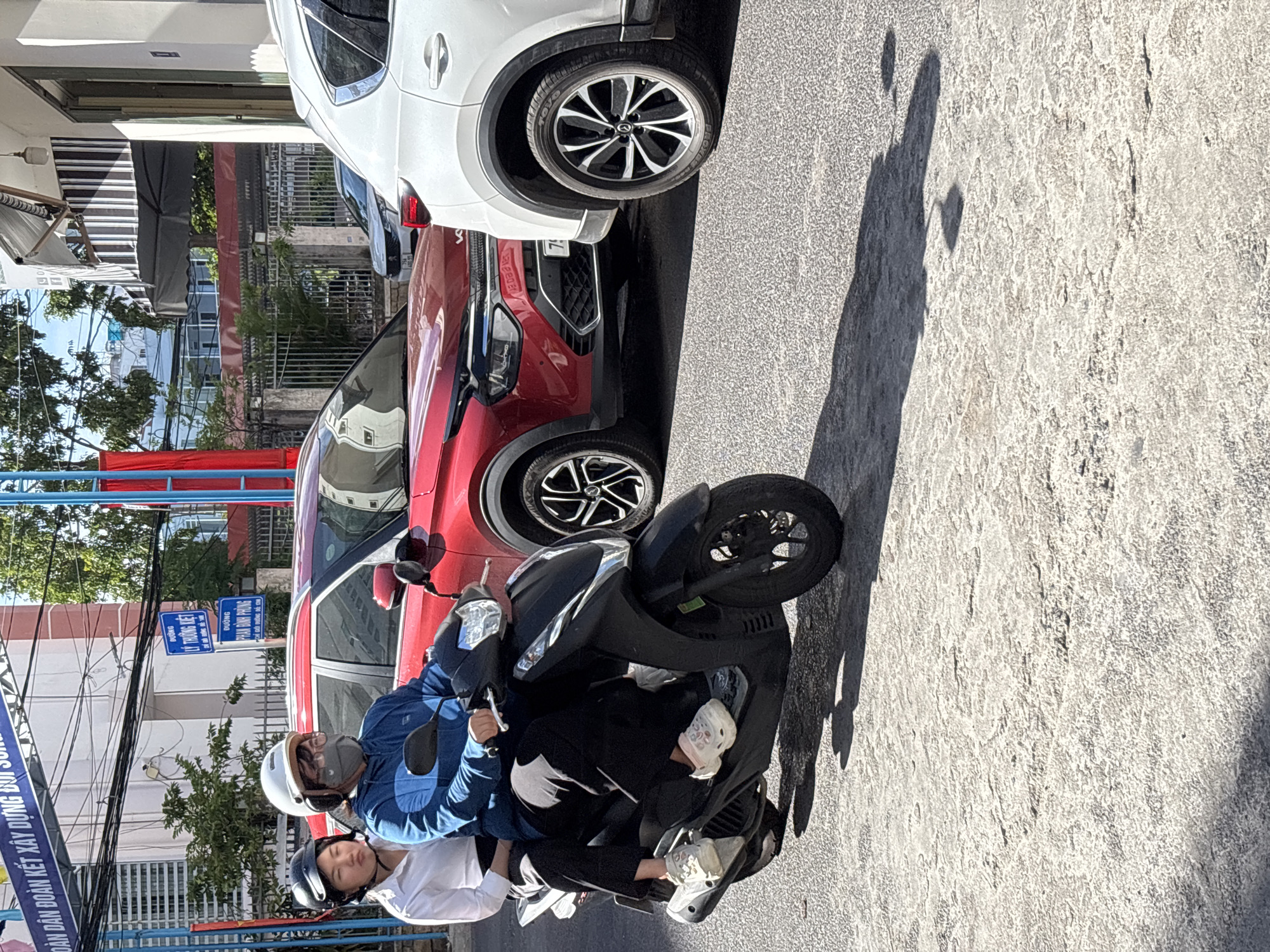 The image depicts a street scene in Nha Trang, Vietnam, on a sunny day. Two individuals are riding a black scooter. The person in front, likely the driver, is wearing a blue jacket, a white helmet, and a gray mask. They are looking forward. The passenger behind them is wearing a white shirt and a black helmet, and appears to be looking down. Both are wearing casual footwear.
In the background, a red SUV is parked or moving slowly, with its front visible. To the right, the rear section of a white SUV is also visible. The street is paved with textured asphalt, and shadows from the vehicles and an overhead structure are cast across it. Above the street, power lines and signs are visible. One sign is blue with white text, indicating street names. The overall environment suggests an urban setting with moderate traffic.