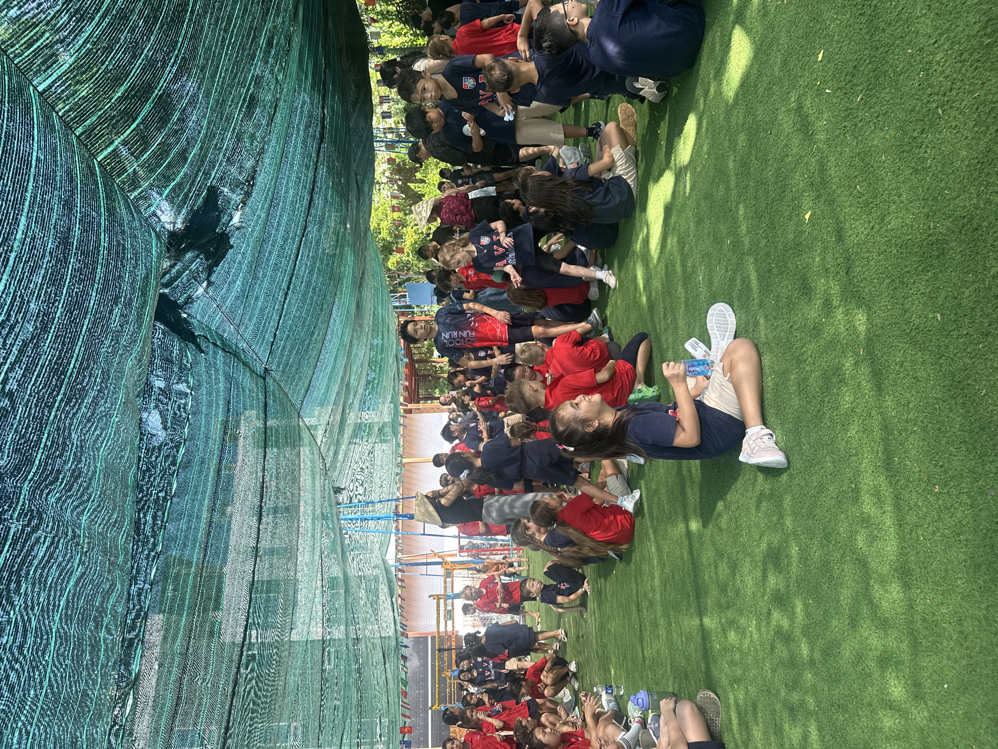 The image depicts a group of children and adults gathered on artificial turf under a green mesh canopy. The setting appears to be an outdoor school or recreational area, likely in Nam Nha Trang Ward, Vietnam, given the presence of colorful flags strung across the background, typical of school events or festivals in the region. The ground is covered in green artificial grass, and the overhead mesh provides shade from the sun, creating dappled light patterns.
In the foreground, several children are seated or kneeling on the turf, some in red t-shirts and others in navy blue. They appear to be resting, talking, or watching something. One child in the center foreground, wearing a navy blue t-shirt and beige shorts, is looking towards the right. To the left, another child in a red shirt is seated with a water bottle.
Further back, a larger crowd of children and some adults are standing and milling about. Many of the children are wearing matching uniforms, predominantly red or navy blue t-shirts with school logos. One adult in the center background is wearing a conical hat, a traditional Vietnamese headwear, suggesting the cultural context of the location. The overall atmosphere seems to be one of a communal gathering, possibly a school event or a sports activity.
In the background, there are structures that resemble a playground or sports field, including a volleyball net and what looks like a slide or climbing frame. The presence of various flags adds a festive and possibly international element to the scene. The trees and foliage visible beyond the canopy indicate an outdoor environment. The scene is well-lit, with natural sunlight filtering through the mesh canopy.