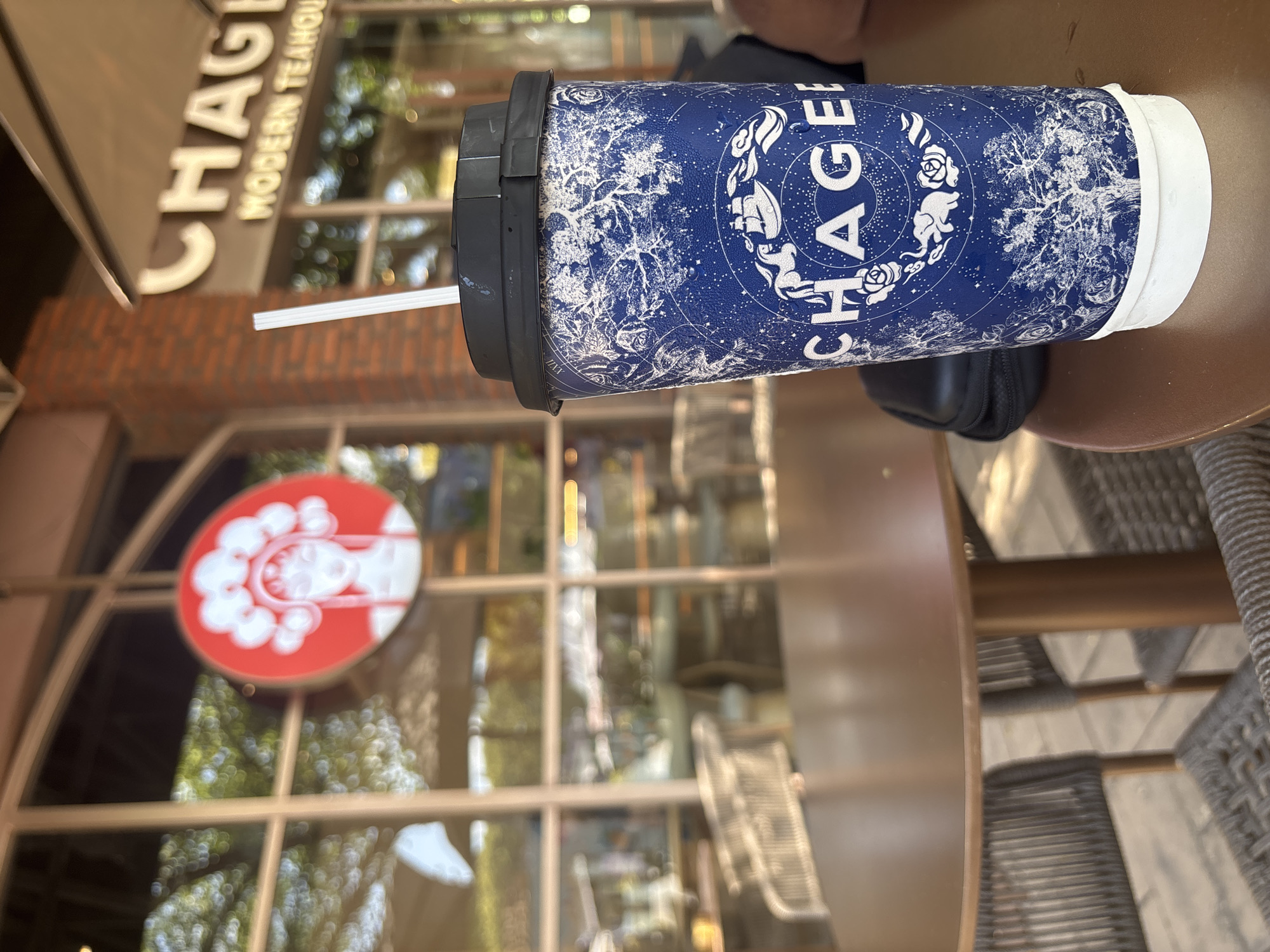 An outdoor seating area at a modern teahouse in Kuta Selatan, Indonesia, is captured in this close-up shot. The focus is on a blue paper to-go cup adorned with white celestial-themed graphics, including a ship and an elephant, and the text "CHAGEE." A white straw protrudes from the black lid. In the background, through large glass windows, a red circular sign featuring a stylized profile of a woman with an elaborate headdress is visible, along with the illuminated text "CHAGEE MODERN TEAHOUSE." The environment includes outdoor tables and chairs, and the general ambiance suggests a bright, sunny day with trees and foliage visible outside.