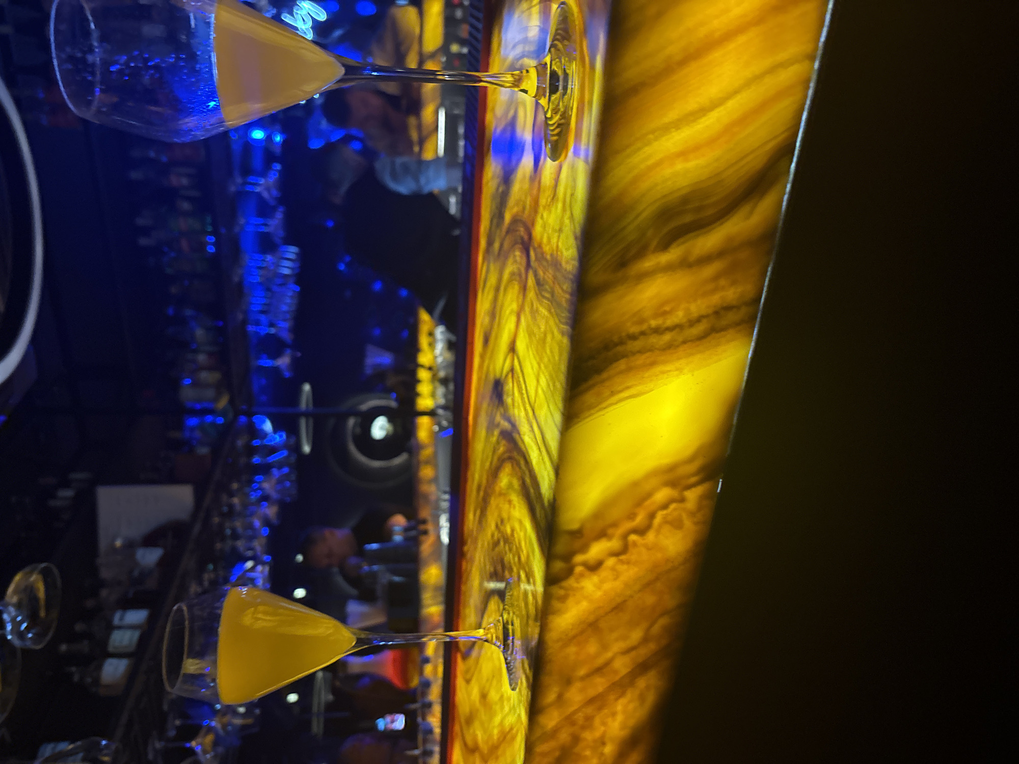 The image depicts a bar scene in Perm, Russia, featuring two champagne flutes filled with a yellow liquid, possibly a mimosa. The flutes are positioned on either side of the frame, reflecting in a glossy surface. The background includes a bar setting with illuminated liquor bottles and blurred figures of people. A prominent feature is the illuminated honey-colored onyx bar top, displaying swirling patterns. The left side of the frame shows a blue-lit area, while the right side transitions into a dark space.