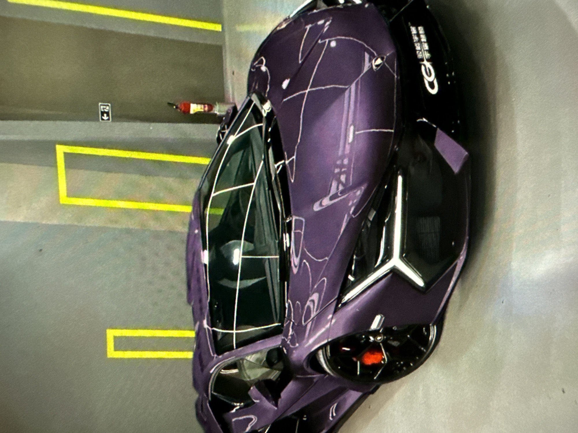 A plum-colored sports car with a glossy finish is parked in a garage. The vehicle is situated within painted yellow parking lines on a light gray surface. An emergency exit sign and a fire extinguisher are visible on the wall behind the car. The location is Unknown, Unknown.