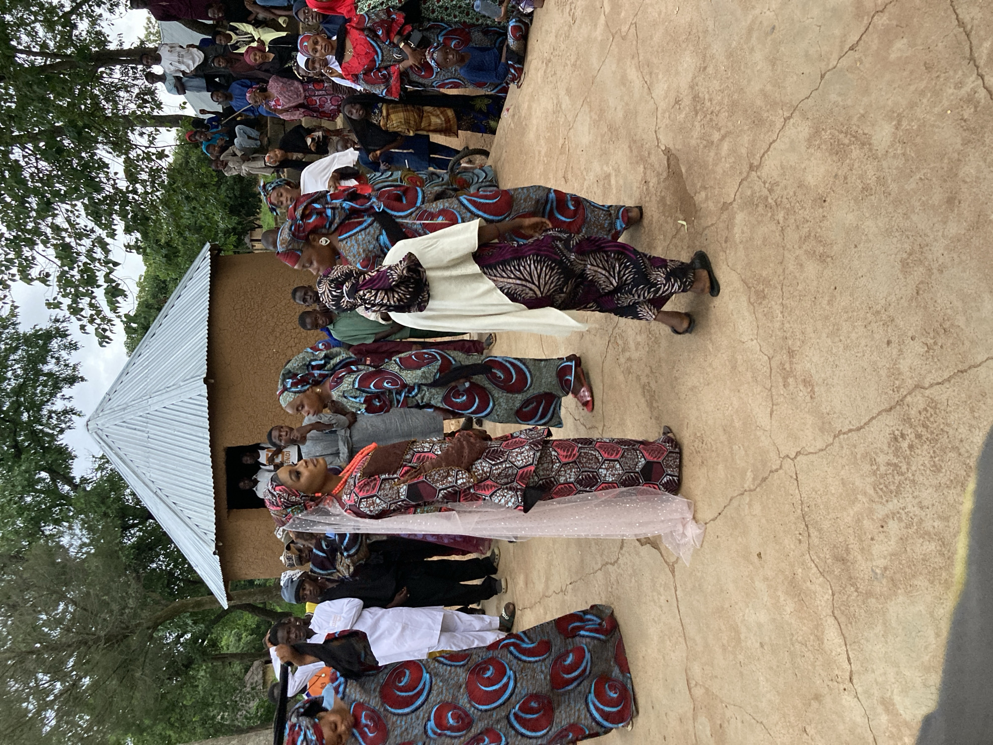 The image depicts an outdoor gathering in Jos, Nigeria, possibly a wedding or a similar celebration. The scene is dominated by people dressed in vibrant, traditional African attire, characterized by bold patterns and bright colors.
In the foreground, a group of women are the central focus. One woman, likely the bride, is dressed in an intricately patterned outfit, a veil, and prominent red beaded jewelry. She is engaged in conversation with another woman in similar elaborate clothing. To the left, another woman in a blue and red patterned dress stands facing away from the camera. Several other women in the foreground are also adorned in colorful, patterned garments.
Behind the foreground figures, a traditional round building with a corrugated metal roof stands prominently. A group of people are gathered around the entrance of this building, some appearing to be guests or attendees. Further into the background, a larger crowd of people are seated and standing, observing the proceedings. The environment appears to be rural or semi-rural, with trees and foliage visible, suggesting an open-air event. The ground is a light brown, cracked earth surface. The overall atmosphere appears to be festive and communal.