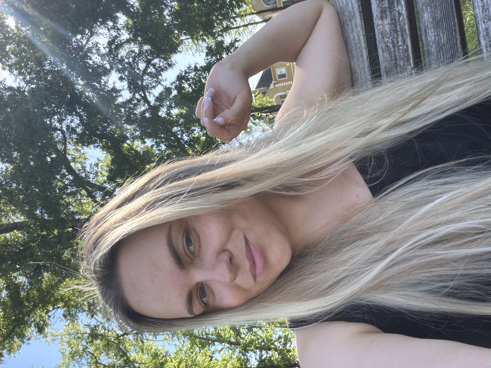 A woman with long, blonde hair is posing for a selfie outdoors on a sunny day. Her face is angled slightly to the right, and she is resting her arm on a wooden bench. The background features trees and a building. The specific location in Votkinsk, Russia, cannot be determined from the image.