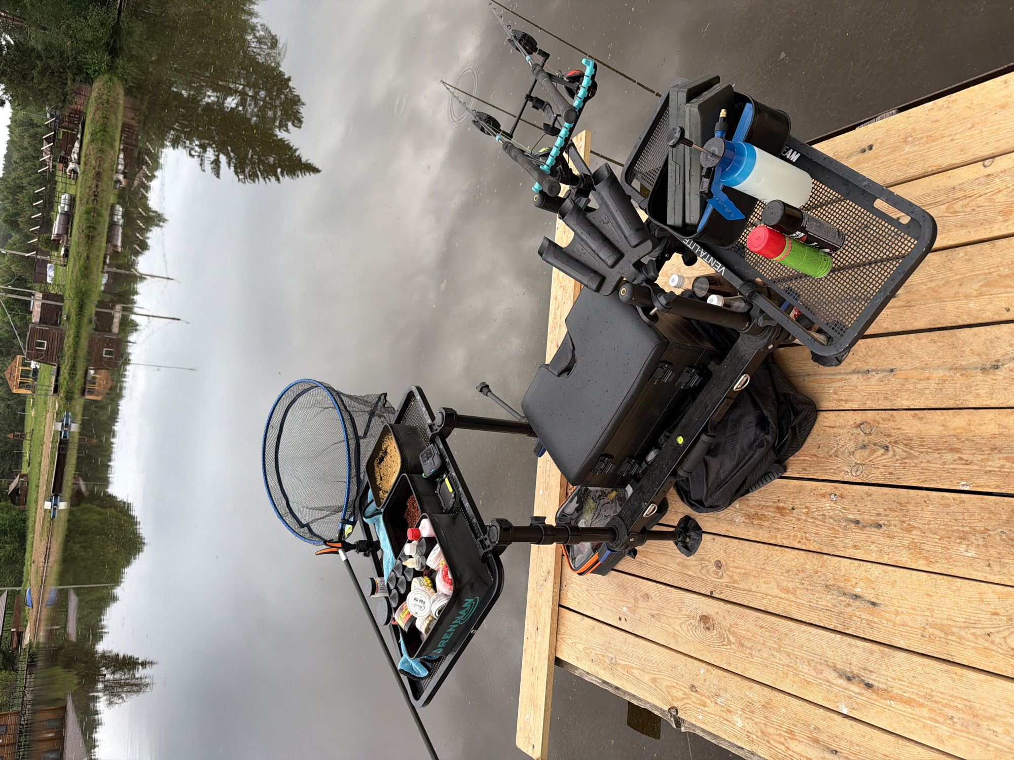 The image shows a fishing setup on a wooden dock, likely in Prikamskii, Russia, given the prompt. The setup includes a fishing seat box with accessories attached. There are two fishing rods with rod rests, a landing net, bait trays, and various containers and bottles. The seat box has a black cover and is placed on a platform with adjustable legs. The bait trays contain various fishing baits and accessories. The background shows a calm body of water with trees and buildings reflected on the surface. The dock is made of light-colored wooden planks. The overall lighting suggests overcast weather.