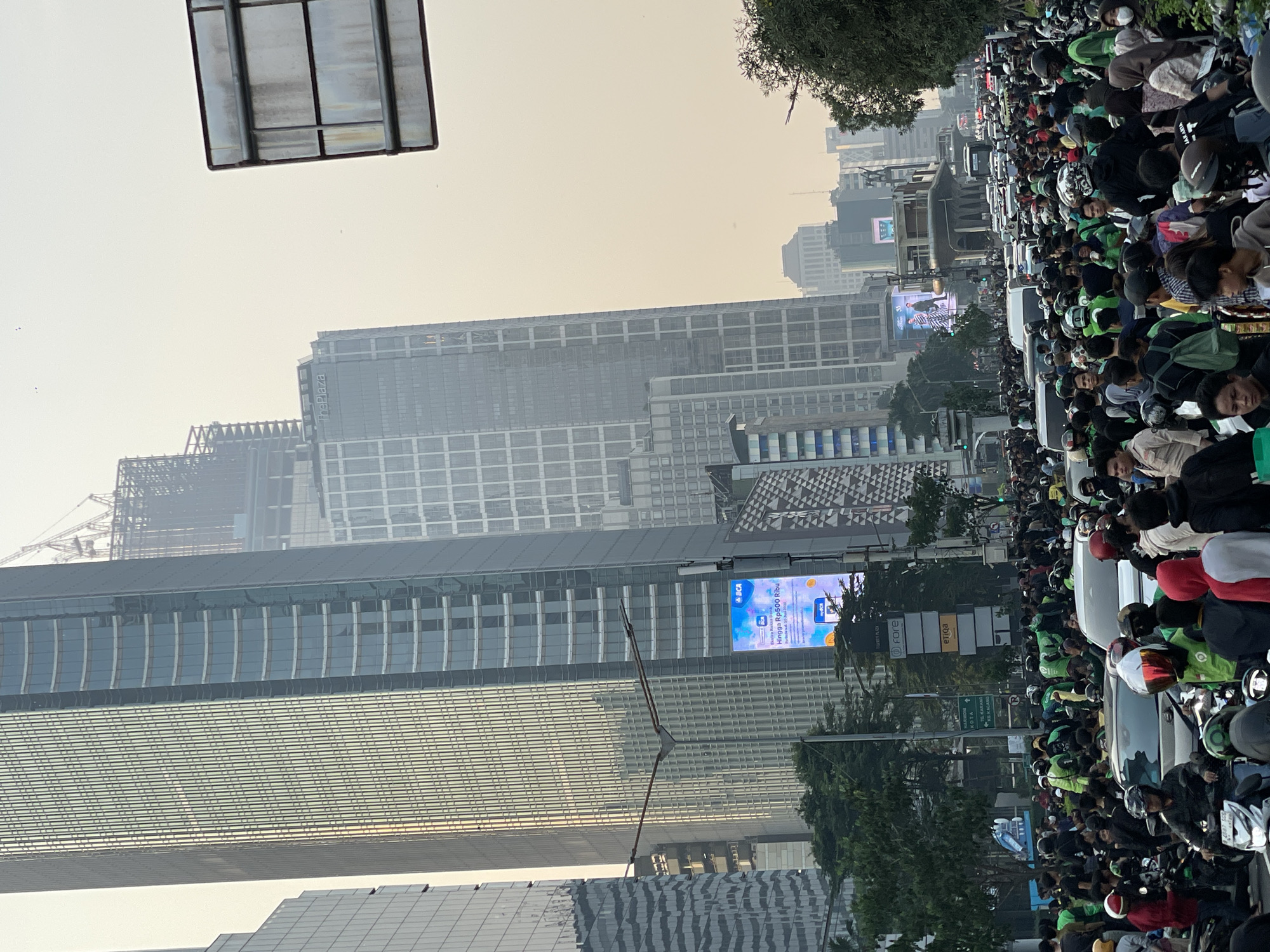 The image depicts a congested street scene in Central Jakarta, Indonesia, with tall modern buildings dominating the skyline. A significant number of people, many wearing green jackets, are gathered around motorcycles and cars, suggesting a traffic jam or a planned gathering. The sky is overcast with a hazy, yellowish hue, indicating possible smog or pollution. Traffic signals and street signs are visible, along with advertisements on digital billboards. The overall impression is one of urban density and a busy, bustling environment.