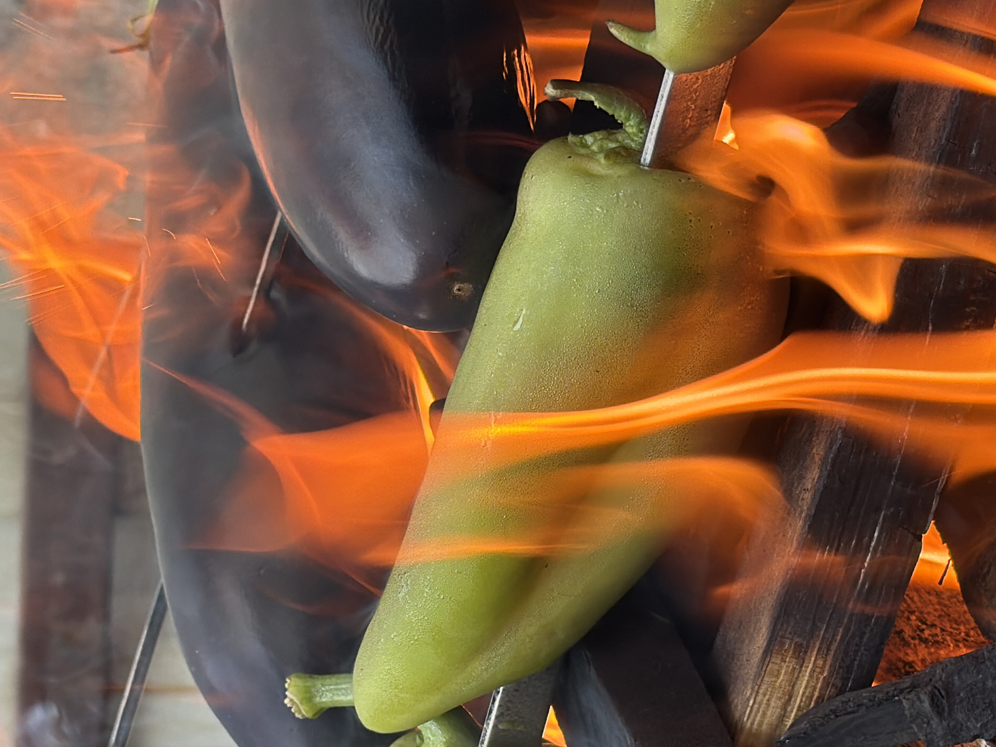 In Tuapse, Russia, a close-up shot captures several vegetables, including eggplants and a green pepper, skewered and being roasted over an open fire. The flames are vibrant orange and yellow, licking around the vegetables and casting a warm glow. The vegetables appear to be on metal skewers, which are resting on a wooden surface that is also being subjected to the heat. The background is out of focus, but hints at an outdoor setting, possibly a barbecue or a cooking event. The image focuses on the textures and colors of the food and the fire, highlighting the cooking process.