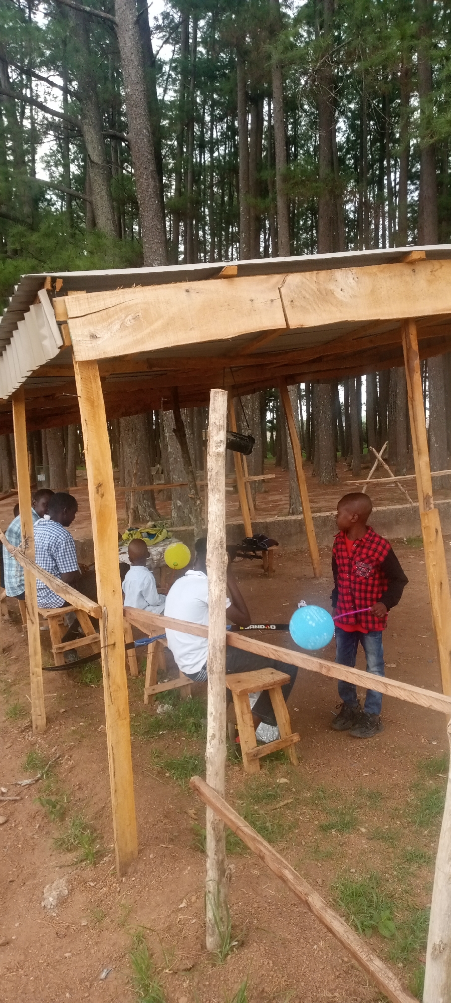 The image shows a group of people gathered under a wooden shelter in what appears to be a wooded area. Tall, thin trees surround the shelter. Inside, several people are sitting on simple wooden stools. One child stands with a blue balloon. Another child has a yellow balloon. There are multiple wooden posts acting as support for the shelter. The ground is reddish-brown dirt with patches of grass. The location is Jos, Nigeria.