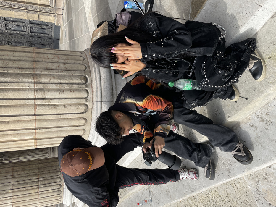 An outdoor scene captured at L'Église de la Madeleine in Paris, France, depicts three individuals on the grand stone steps beneath classical fluted columns. To the left, a standing person wearing a brown baseball cap and black track suit with red stripes is bent over, looking down. In the center, a male individual with dark, curly hair is seated, holding a black camera with a large lens, seemingly adjusting settings or reviewing images. He wears a jacket with an abstract orange and black pattern, and dark jeans. To the right, a female with long, dark hair and bangs is also seated, clad in a studded black denim jacket and ripped black jeans with lacing details. She holds both hands up near her face, fingers splayed, revealing pastel-colored fingernails. Two plastic water bottles, one with a red cap and one with a green cap, are visible on the steps near the individuals. In the background, beyond the columns, the intricately carved stone double doors of the church are visible.
