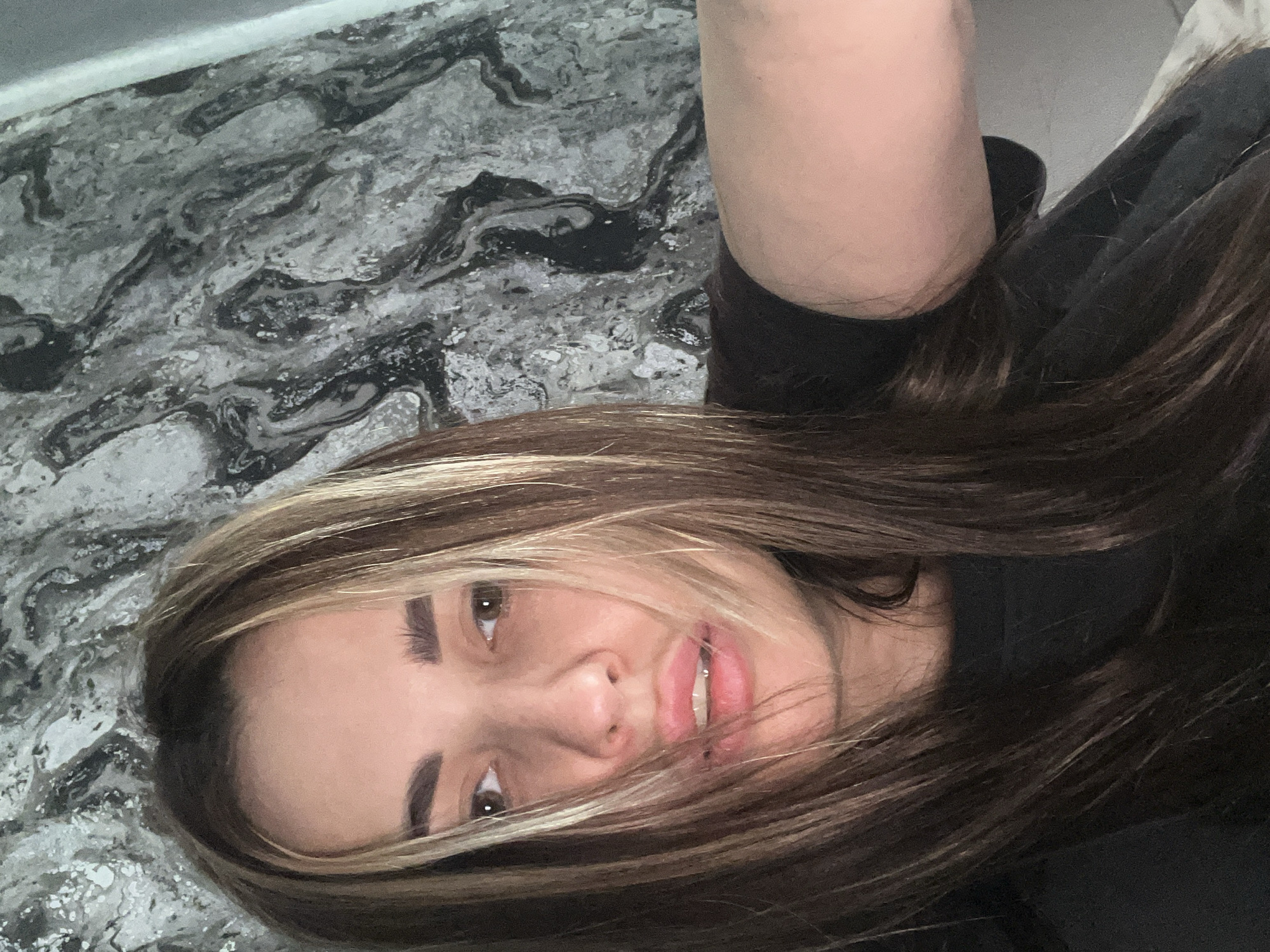 A person with long brown and blonde hair is lying on a gray and white marbled surface. The person is wearing a dark-colored garment. The location is Unknown, Unknown.