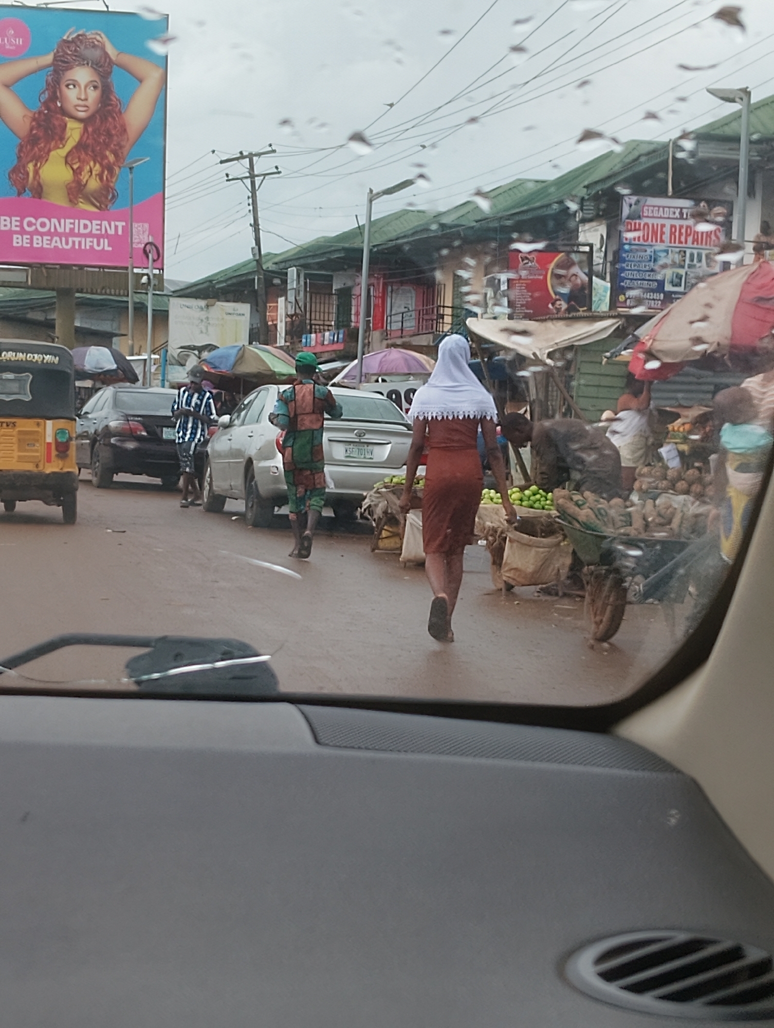 This is a street-level, eye-level, medium shot image taken from inside a vehicle, looking out at a busy marketplace scene. The image captures a somewhat overcast sky with visible raindrops on the windshield, suggesting it might be raining or has recently rained. The ground is wet and reflects the surrounding environment.
In the foreground, the interior of a car is visible, including the dashboard with a speaker grille and a partially obscured windshield wiper. The main focus of the image is the street scene.
Several people are present. A woman wearing a white hijab and a brown dress is walking away from the camera. To her left, a man in a camouflage-patterned uniform with a green cap is also walking, his back to the camera. Further down the street, another person in a black and white striped shirt and shorts is standing, seemingly interacting with someone or looking at something. Other individuals are visible in the background, including a person wearing a bright green cap and a man in what appears to be a blue and white striped shirt and shorts.
A variety of vehicles are present, including a yellow and black tricycle in the left of the frame and several cars. One car, a silver sedan, has a license plate visible that reads "KSF 701 HV".
The environment appears to be a bustling market with numerous stalls set up. Colorful umbrellas and awnings are visible over some stalls, providing shade. Produce, including what looks like green fruits and root vegetables, are displayed in carts and on the ground. Signs are visible on buildings, including one advertising "SEGADEX TECHNOLOGY PHONE REPAIRS" with smaller text listing services like "FIXING", "REPAIRS", "UNLOCKING", and "FLASHING". Another large billboard on the left features an advertisement with the text "BE CONFIDENT BE BEAUTIFUL" and an image of a woman.
The overall impression is of a dynamic, everyday scene in a commercial area, likely Aketu-Oja, Nigeria, given the context.