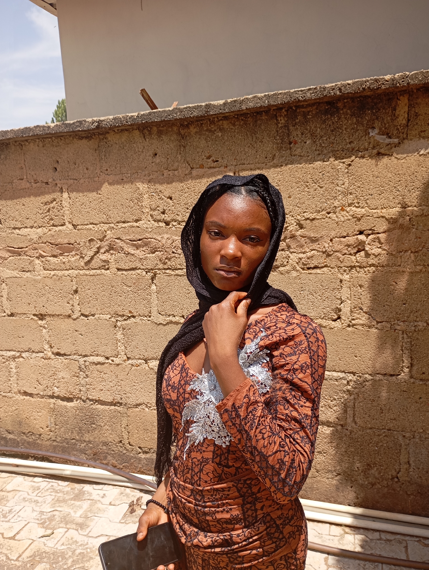 A young, dark-skinned woman stands outdoors, posing for a photograph. She is in front of a weathered, brick wall with a concrete border. The woman is wearing a patterned orange and black dress with silver embellishments on the chest, and a black headscarf. She is holding a black smartphone in her left hand and has her right hand near her neck, adjusting her headscarf or posing. The location appears to be in Jos, Nigeria based on the style of dress and wall construction. The light suggests it is daytime. A white pipe runs along the bottom of the wall, near the ground. The bricks of the wall are uneven and bear the marks of age. The background is partly shaded.