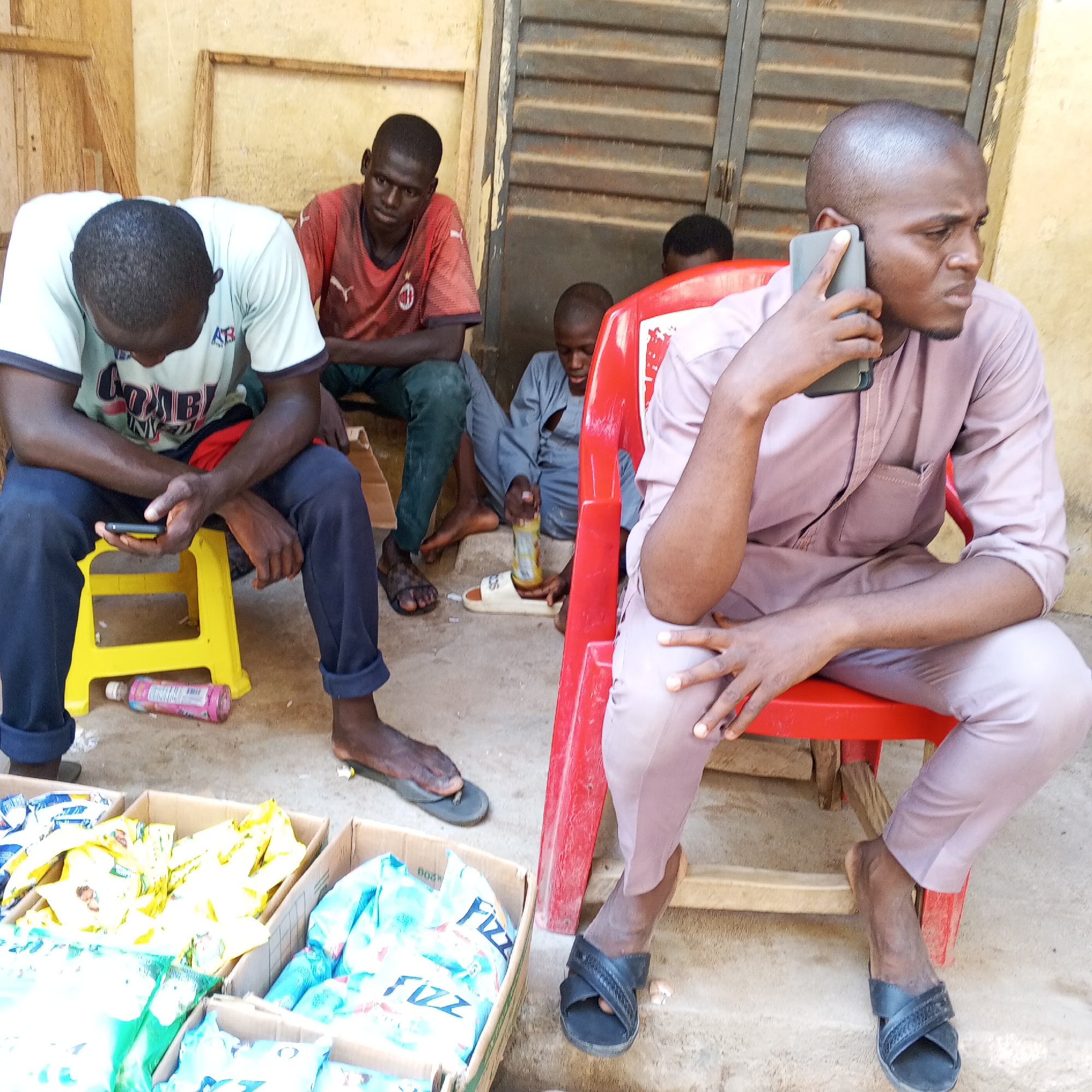 The image depicts a group of young men in a semi-outdoor setting that appears to be a marketplace or vendor stall in Gombe, Nigeria. In the foreground, a man in a light purple traditional outfit is seated on a red plastic chair, holding a smartphone to his ear. To his left, another man in a light green t-shirt and dark pants is seated on a yellow plastic stool, looking down at a smartphone in his hand. Behind these two men, two other individuals are visible. One, wearing a red t-shirt, is seated with his arms crossed. Another, in a grey garment, is seated further back. The ground is dusty, and several cardboard boxes filled with packaged goods, possibly snacks or drinks, are arranged in front of the seated individuals. A pink plastic bottle lies on the ground near the yellow stool. The background shows a rough wall and a corrugated metal structure, suggesting a simple, possibly temporary, commercial space. The lighting suggests it is daytime.