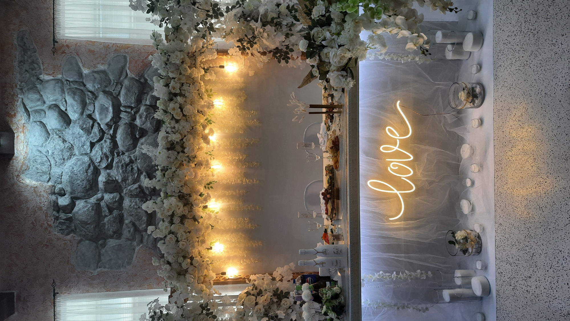 The image depicts a wedding reception table set up in a venue in the village of Zhodziškava, Belarus. The table is adorned with white floral arrangements, string lights, and a neon "love" sign. Two champagne bottles and a cheese board are visible on the table, suggesting a celebration. The backdrop features a textured wall with a stone accent. The overall atmosphere is festive and romantic, indicative of a wedding ceremony or reception.