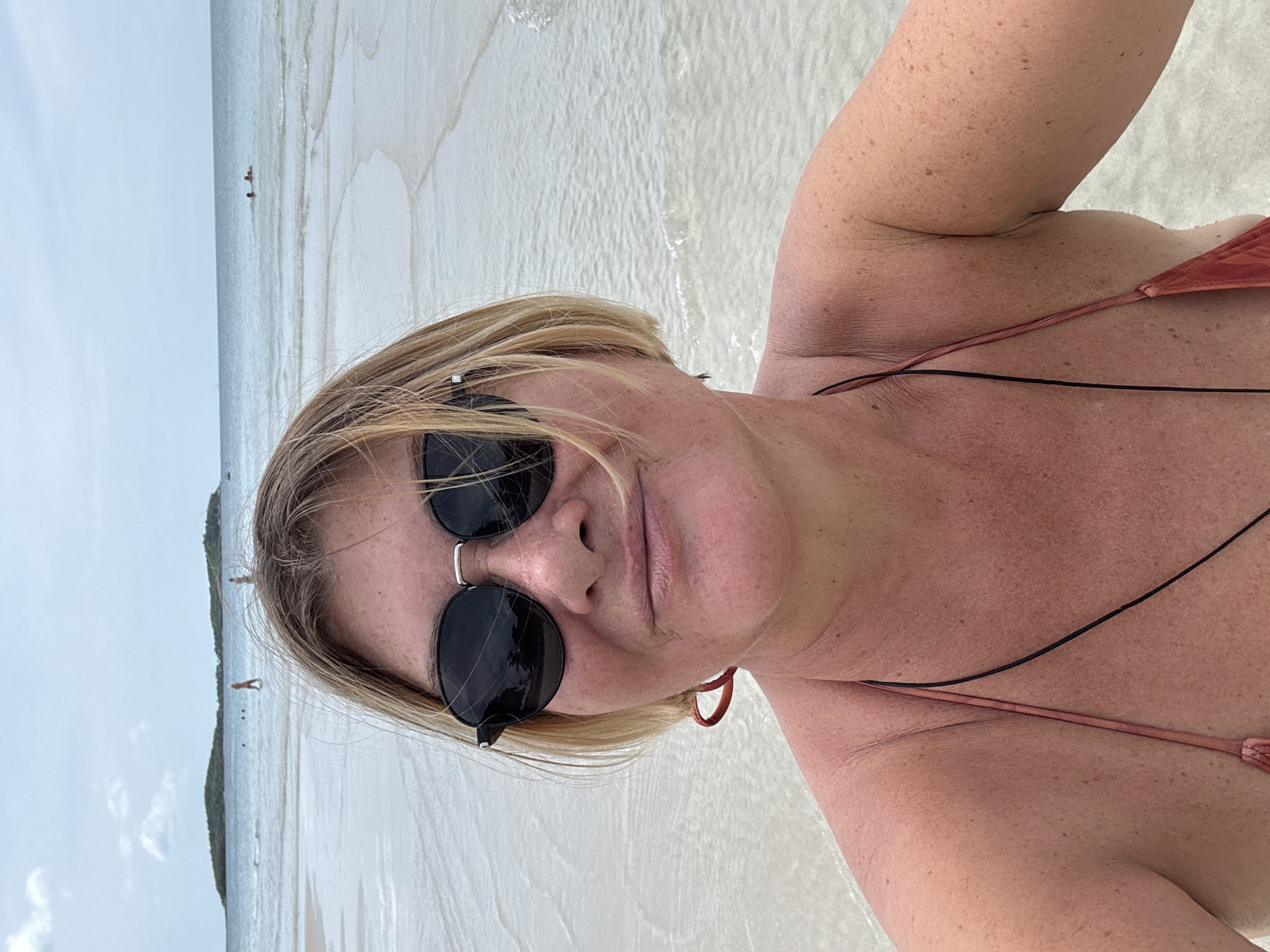 A medium shot selfie shows a woman with short blonde hair and black sunglasses posing on a beach. She is wearing a coral-colored bikini and appears to have sunburn on her shoulders and chest. The background consists of a shallow, clear ocean with gentle waves, a sandy shoreline, and a hazy sky. In the distance, two islands are visible, with a few small figures in the water. The scene is set in Baan Chaweng, Thailand, a tropical coastal town.