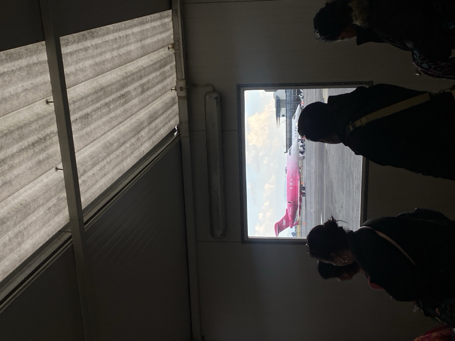 From an airport terminal window, a bright pink Viva Aerobus airplane sits on the tarmac. Several people stand, looking out at the plane. Inside the terminal, corrugated metal and a fluorescent light are visible on the ceiling. The scene is shadowed, contrasting with the bright view of the airplane outside.