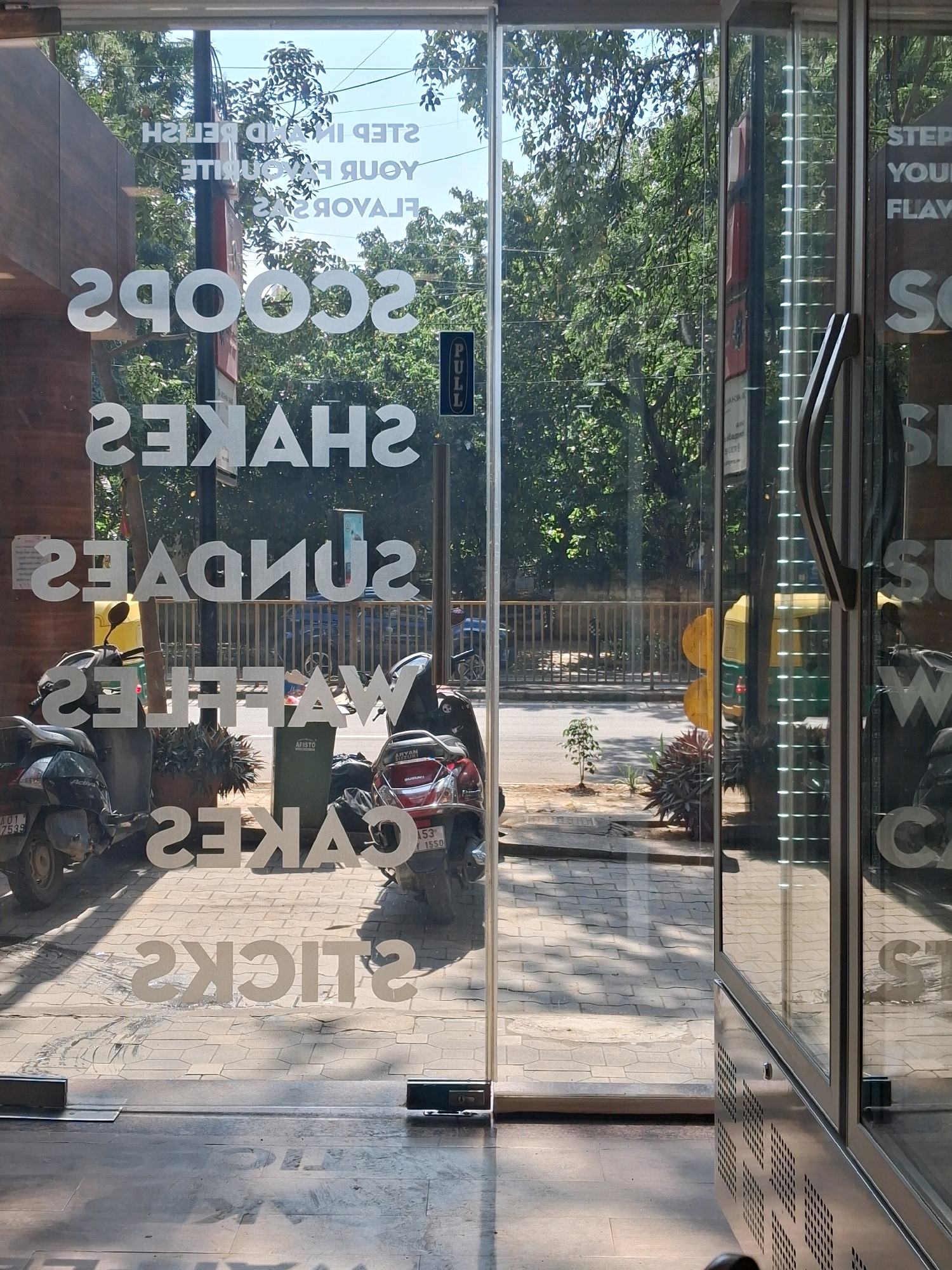 This is a daytime, outdoor, eye-level shot of a storefront glass door, taken from a slightly low angle, showing a street view of Bengaluru, India. The glass reflects the sky, trees, and street with vehicles. The storefront displays a variety of words in white lettering, oriented to be read from outside: "SCOOPS", "SHAKES", "SUNDAES", "WAFFLES", and "CAKES", with "STICKS" visible near the bottom. Above these, also in white lettering, are the words "STEP IN AND RELISH YOUR FAVORITE FLAVORS". A "PULL" sign is affixed to the right side of the glass. Outside the storefront, two scooters are parked. The foreground shows a tiled pedestrian walkway with shadows. A metal trash bin is visible between the scooters. Beyond the walkway, a street with traffic and trees is visible.