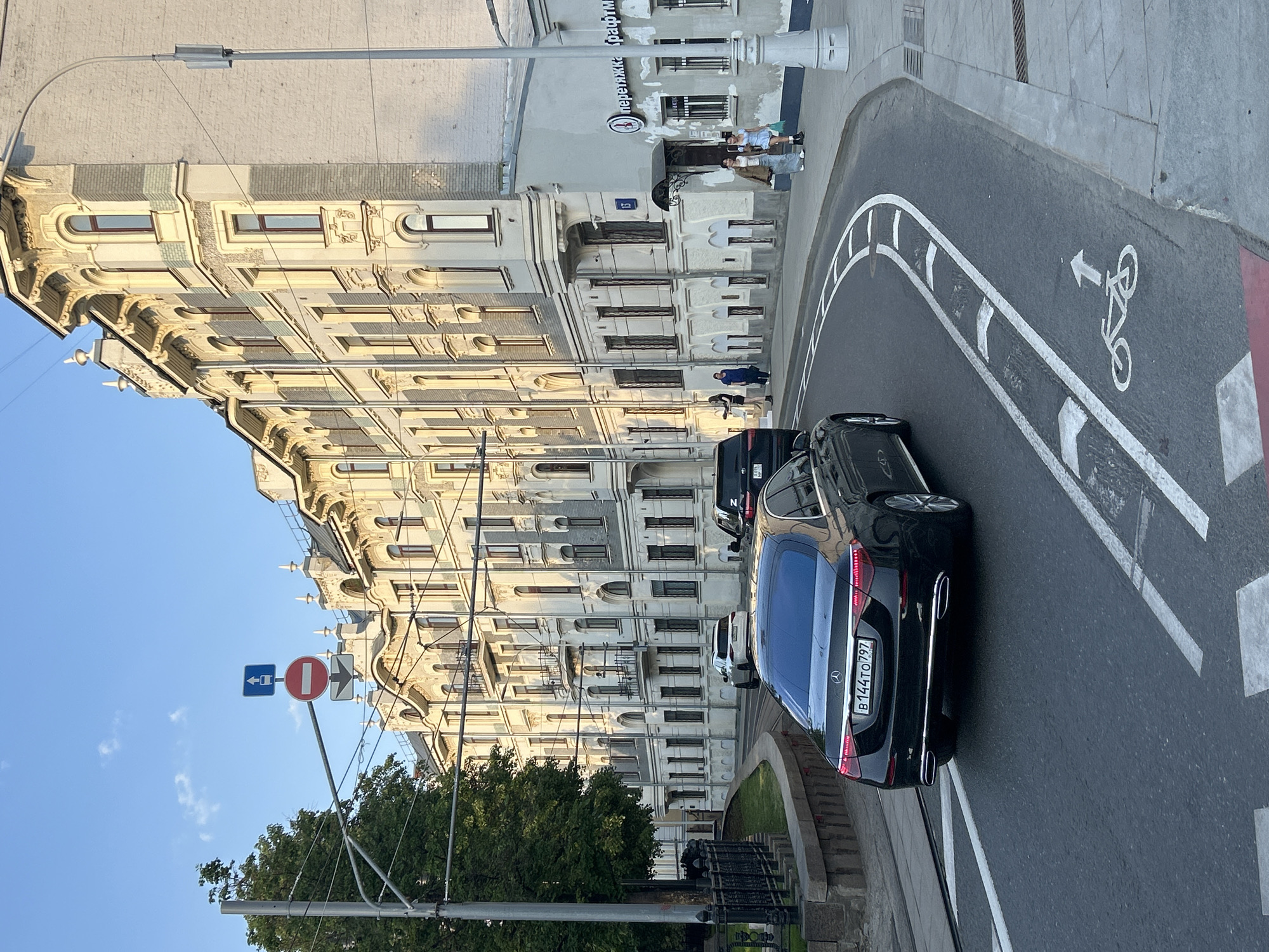 A street scene shows a dark-colored Mercedes sedan navigating a bend in the road. The car's license plate is clearly visible, reading "B 144 TO 797". The road has a designated bike lane, marked with a bicycle symbol and an arrow indicating the direction of travel. In the background, there is a tall, ornate building with multiple windows and detailed architectural features. A street sign is visible, containing traffic regulations. The sky is a clear blue. There are some people visible on the sidewalk in front of the building.
