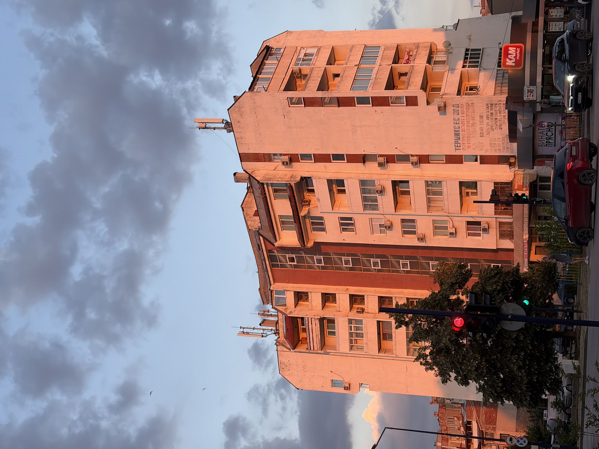 This media file captures a street scene bathed in the warm glow of sunset. The primary subject is a tall, multi-story building with a textured, light-colored facade, illuminated by the orange hues of the setting sun. The building features numerous windows, some reflecting the sky and others showing the interior. Balconies with red railings are visible on some floors. Several air conditioning units are mounted on the exterior walls. Atop the building, antennas and a small structure are present.
To the right of the building, a "KAM market" sign with orange lettering is prominently displayed. Below it, faded text in Cyrillic script appears on the building's wall, possibly an advertisement or company information. A traffic light stands in the foreground, showing a red light, and a lush green tree partially obscures it. Several cars are parked or driving on the street, including a dark-colored car and a red car. The sky is a mix of blue and dramatic, dark clouds, suggesting an approaching evening or a change in weather. The overall impression is one of urban life at dusk, with commercial activity and everyday structures forming the backdrop. The time of day is clearly indicated by the sunset lighting and the traffic light status. No people are clearly distinguishable in the image.