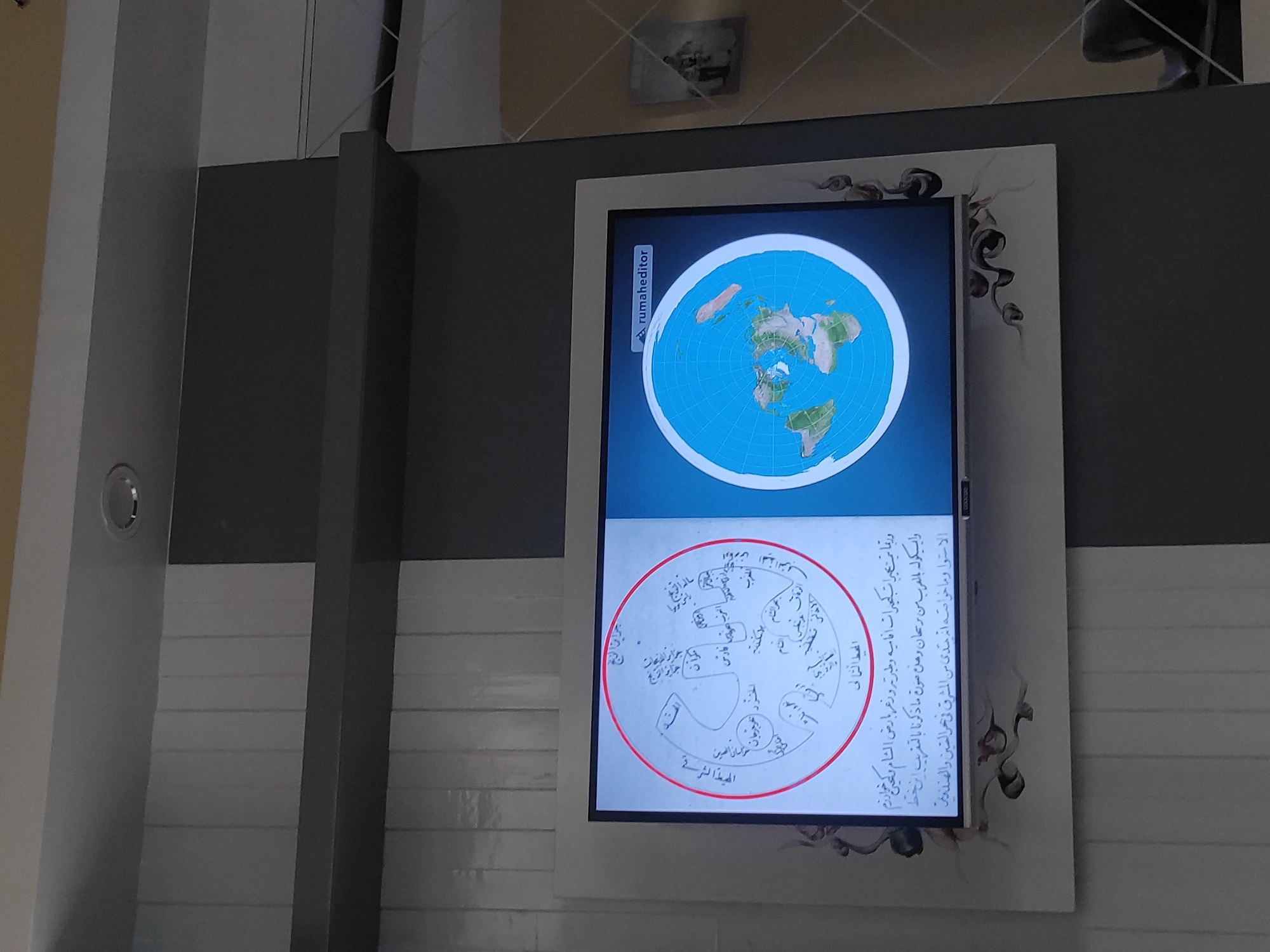 The image shows a television screen displaying two maps. The left map is an ancient circular diagram of the world, surrounded by red ink. The right map depicts a flat Earth projection with continents. The television is mounted on a wall with a grey and white striped pattern. A watermark in the top right corner of the screen reads "rumaheditor". The image quality is moderate, with some pixelation and reflections. The scene appears to be an indoor setting.