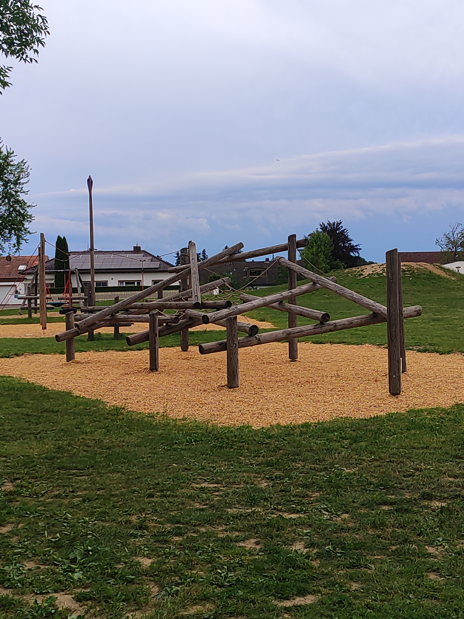 The image depicts a playground obstacle course constructed from natural wooden logs, situated in the municipality of Diedorf, Germany. The obstacle features a series of interconnected logs at various angles, forming a climbing and balancing structure. The ground beneath and around the obstacle is covered with a layer of light brown wood chips, transitioning into a grassy area. In the background, a residential building with a white facade and a dark roof is visible, equipped with solar panels. Trees and a slight incline covered in grass and sand are also present in the surrounding environment. The sky is overcast with visible clouds.