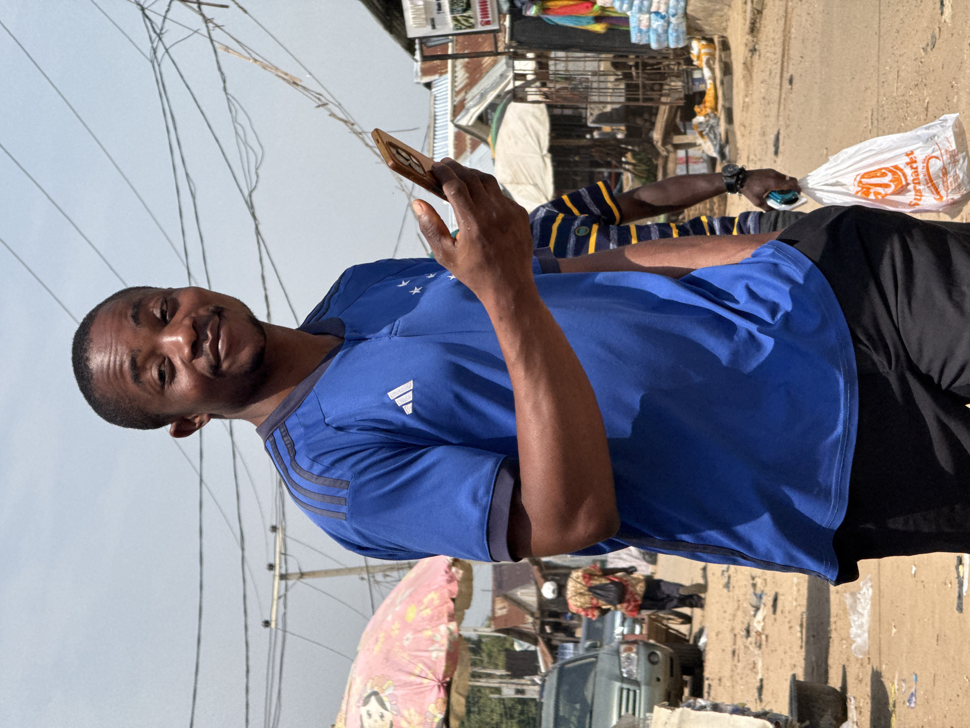 In Abuja, Nigeria, a Black man in his late twenties or early thirties, wearing a royal blue Adidas soccer jersey with white stripes on the sleeves and collar, black athletic pants, and white socks with black athletic shoes, walks down a dusty street. He is holding a gold-colored smartphone in his right hand, angled upwards as if taking a selfie or recording a video. His left hand is in his pocket. He has a slight smile and is looking towards the camera. Behind him, a woman with a white headscarf and a patterned shirt with a black backpack is visible further down the street, walking away from the camera. Another person in a striped blue and yellow shirt is also visible in the background. The street is lined with market stalls, some with corrugated metal roofs, and appears to be a bustling commercial area with various goods displayed for sale, including colorful textiles. Overhead, a complex network of electrical wires crisscrosses the sky, supported by wooden poles. The ground is covered in loose dirt and debris. The sky is clear and bright, indicating a sunny day.